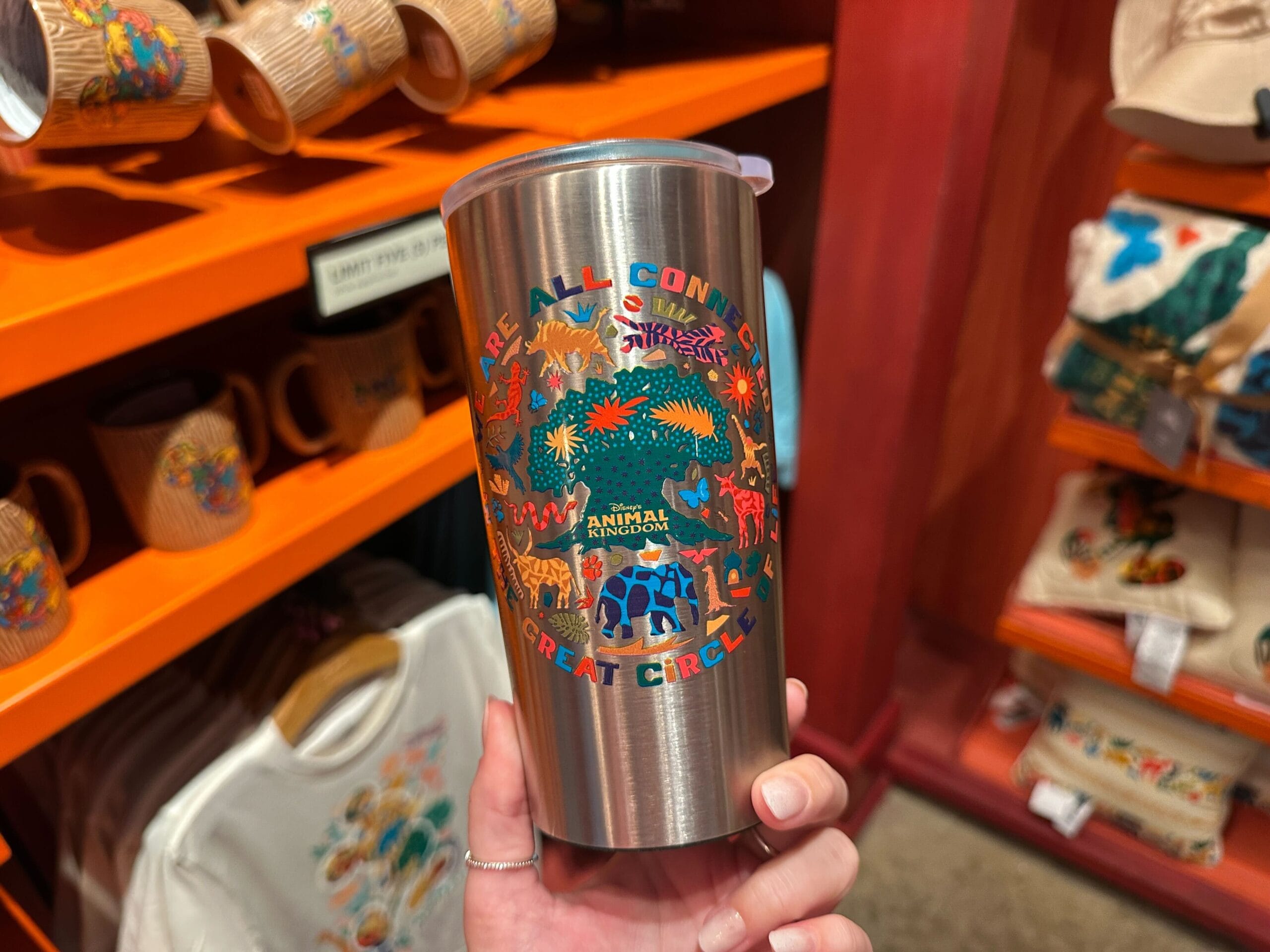 A hand holds a stainless steel tumbler amid colorful Animal Kingdom-themed Disney merchandise in a park gift shop.