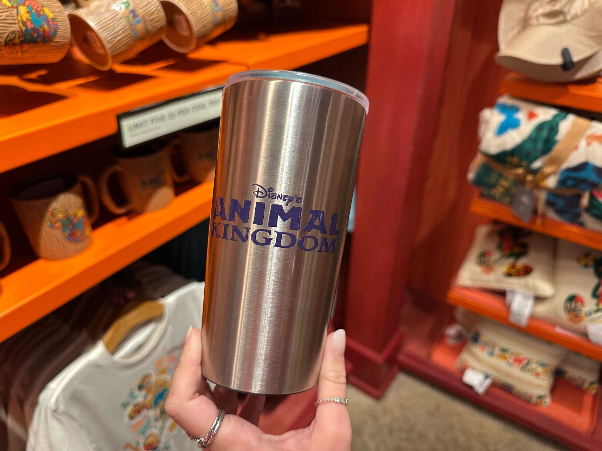 A hand holds a silver Animal Kingdom tumbler inside a shop stocked with themed Circle of Life mugs, shirts, and hats—no castle in view.