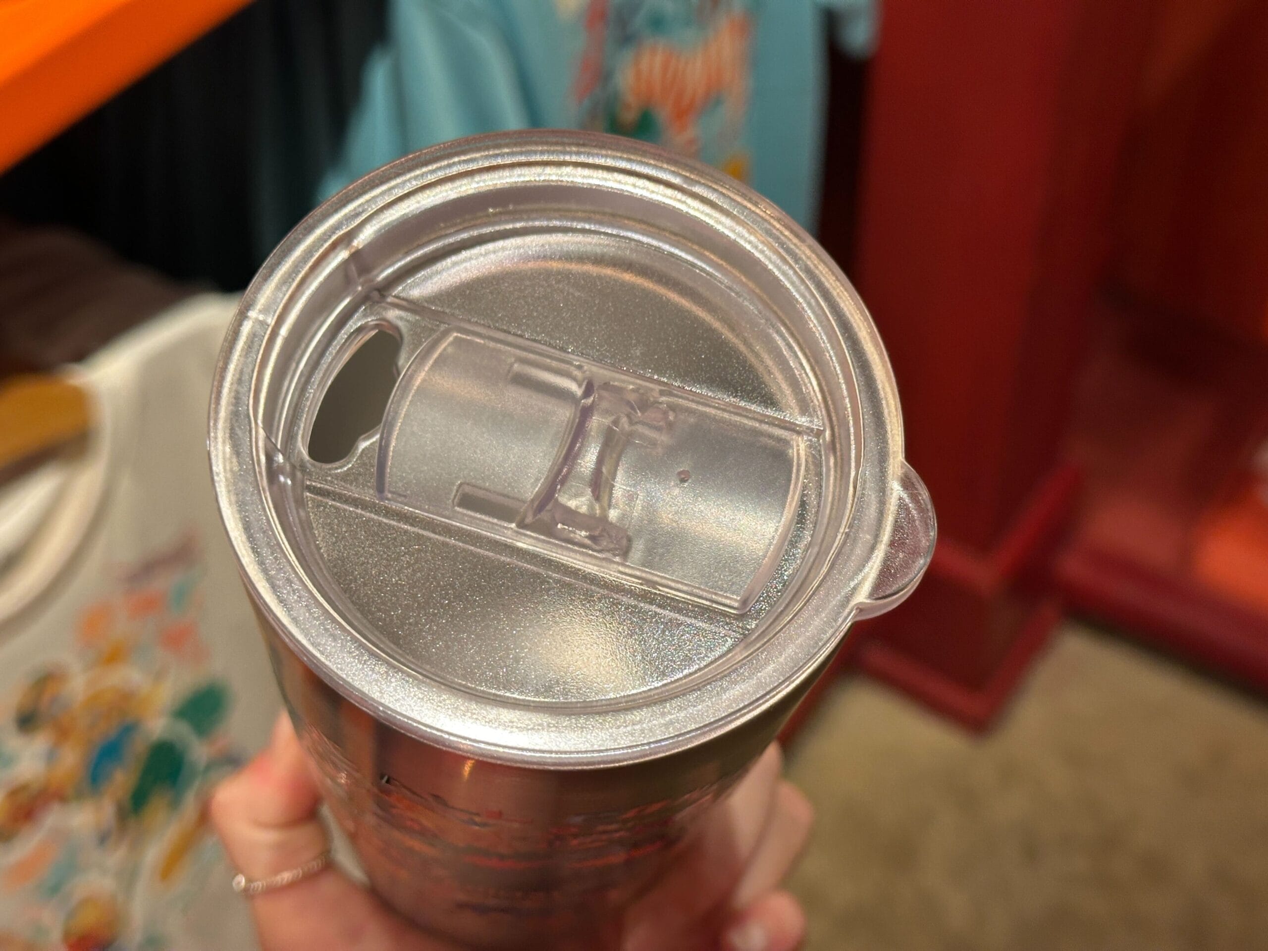 A close-up of a Disney Parks clear tumbler lid with sliding cover, held indoors—reminiscent of souvenir cups from park shops.