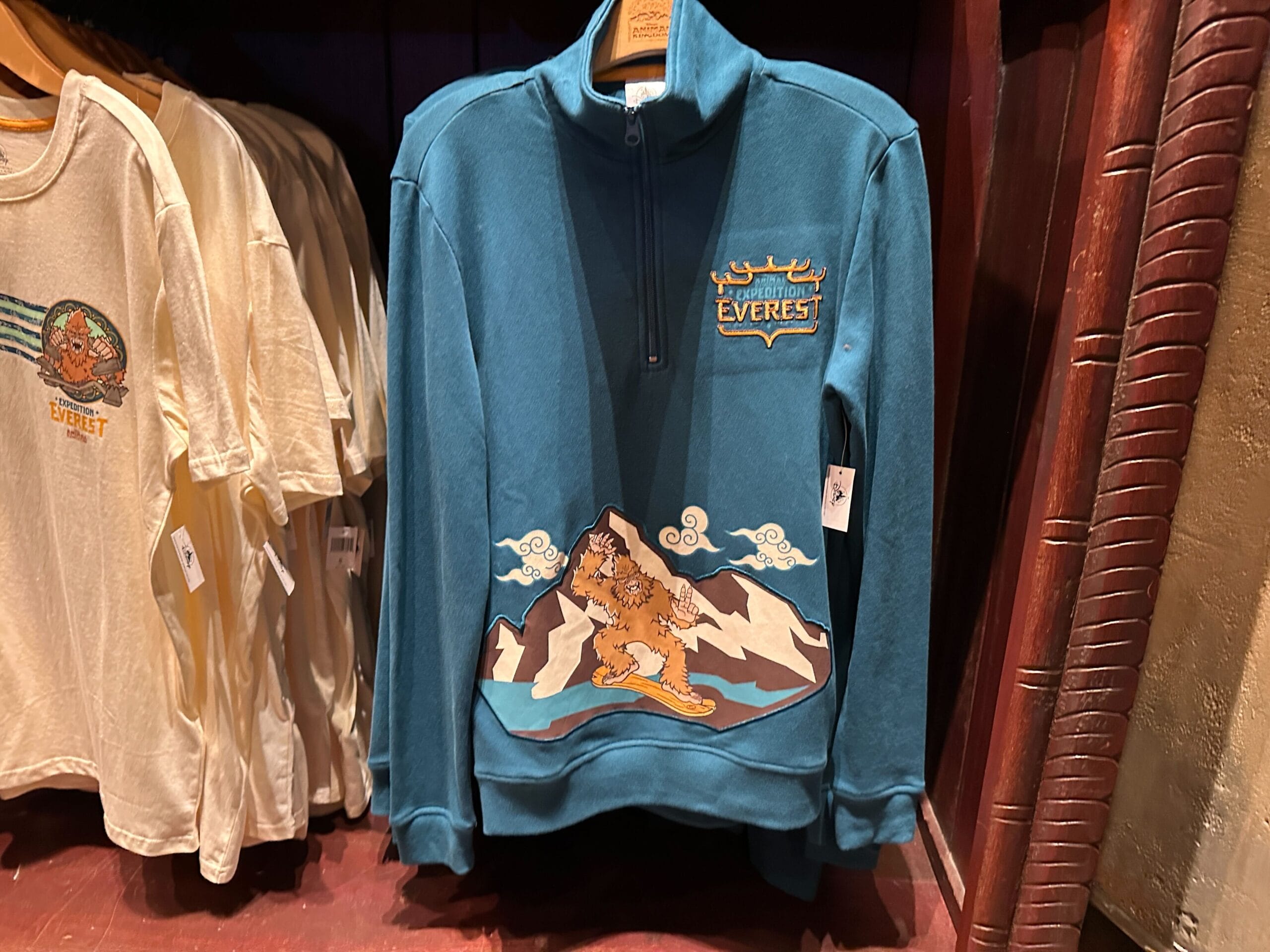 Teal "Expedition Everest" pullover featuring a yeti, mountains, and clouds, seen on a Disney World shop rack—no castle nearby.