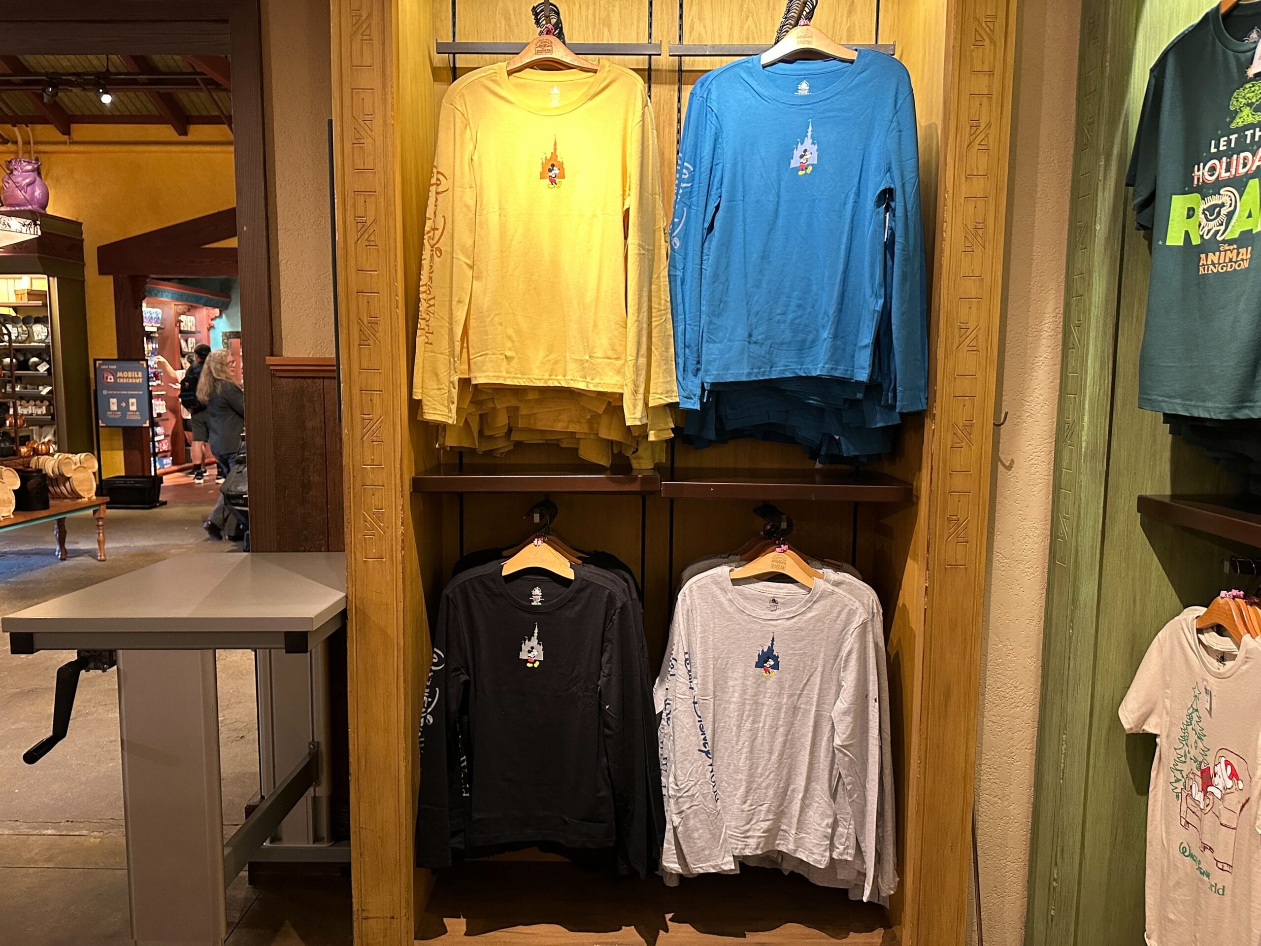 Four long-sleeve shirts and a plush Mickey Mouse Sherpa Hoodie are displayed in a Disney Parks shop, perfect for cool days.