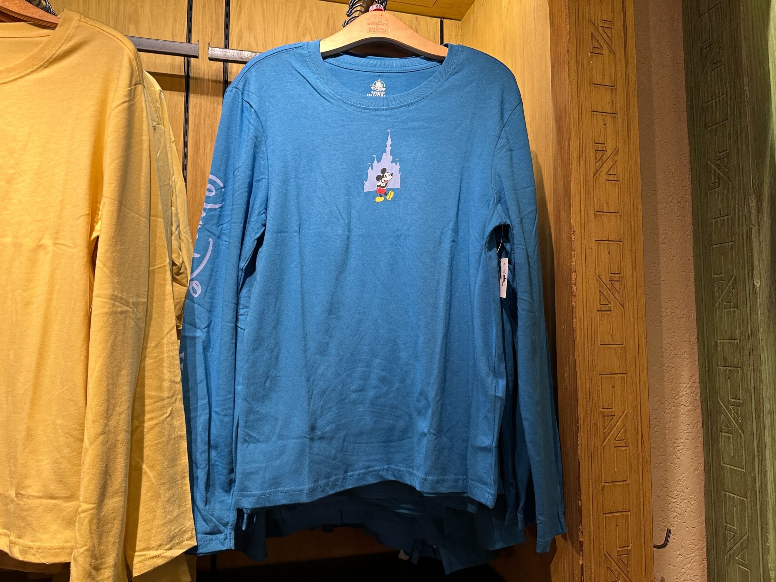 Blue long-sleeve tee with small Mickey Mouse and a castle graphic on the chest, resembling Cinderella Castle at Disney World, hanging on a rack.