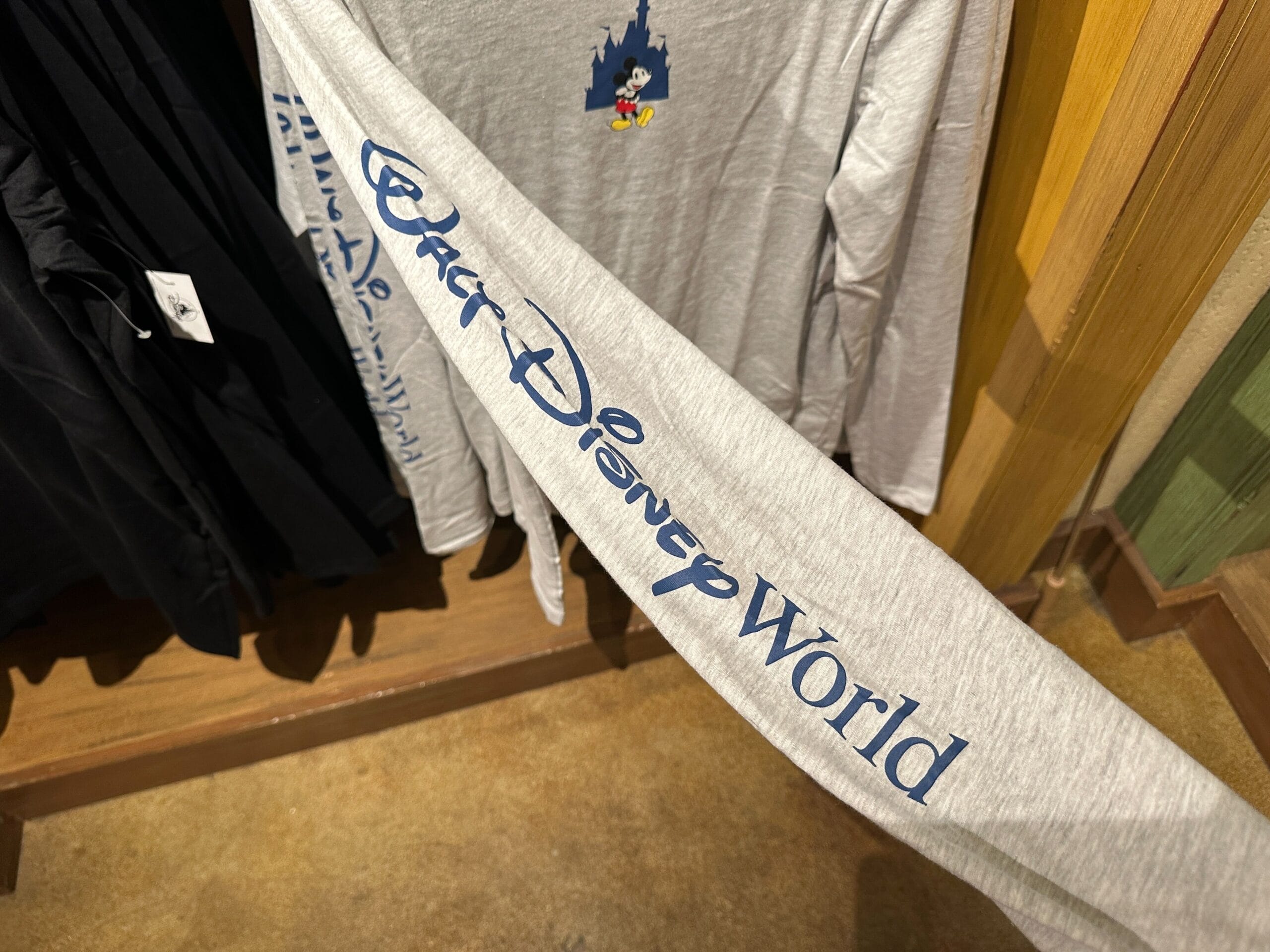 A gray long-sleeve shirt featuring "Walt Disney World" on the sleeve, displayed near plush Mickey Mouse Sherpa Hoodies in park shop.