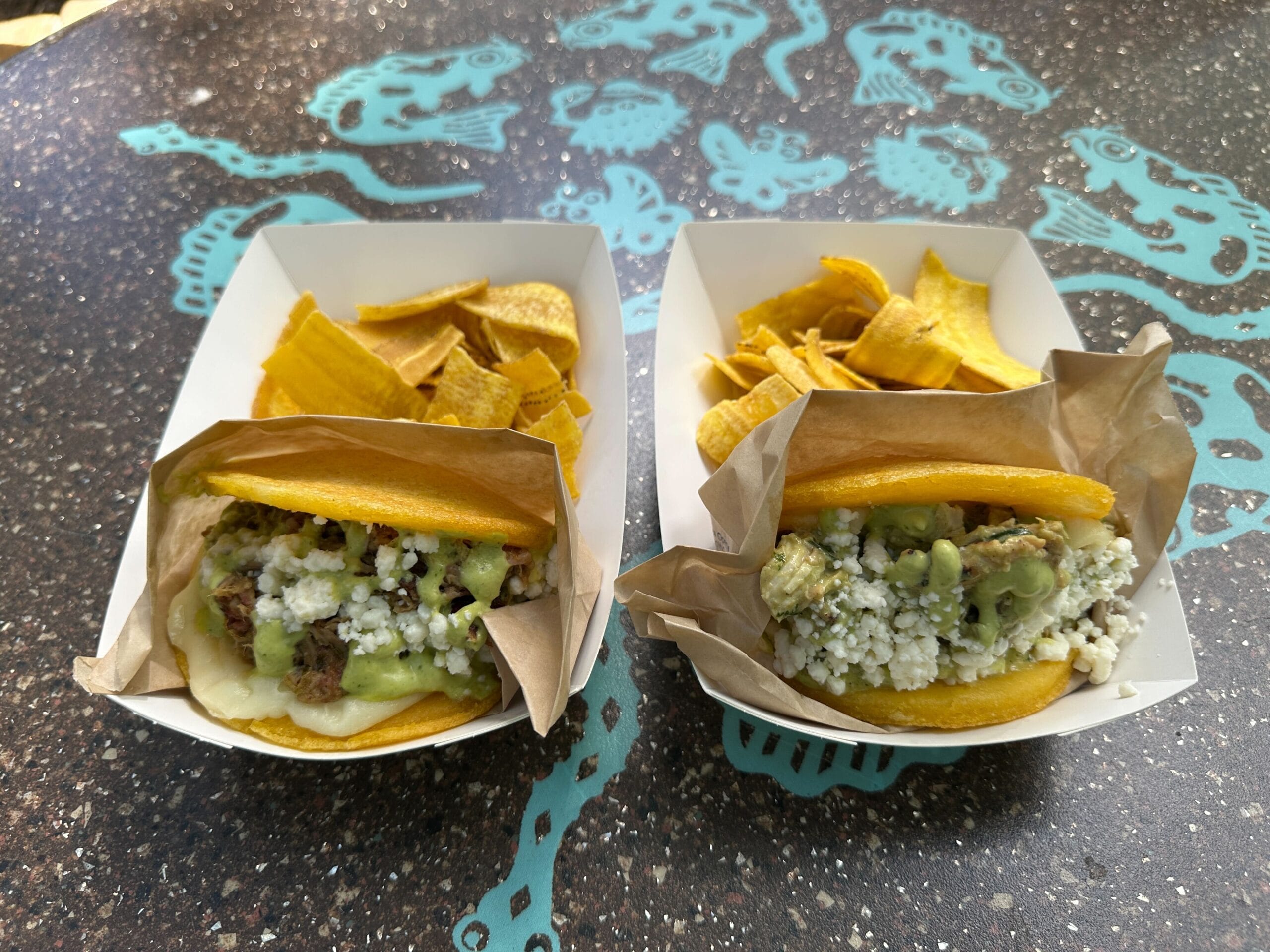Two arepas with cheese, avocado, and meat in paper trays with plantain chips at Terra Treats, a Walt Disney World snack spot.