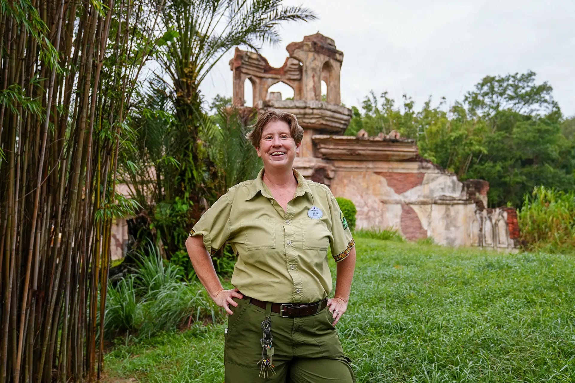 A smiling woman in a green uniform stands outside, posing for TIME for Kids with stone ruins and greenery like Disney park scenery.