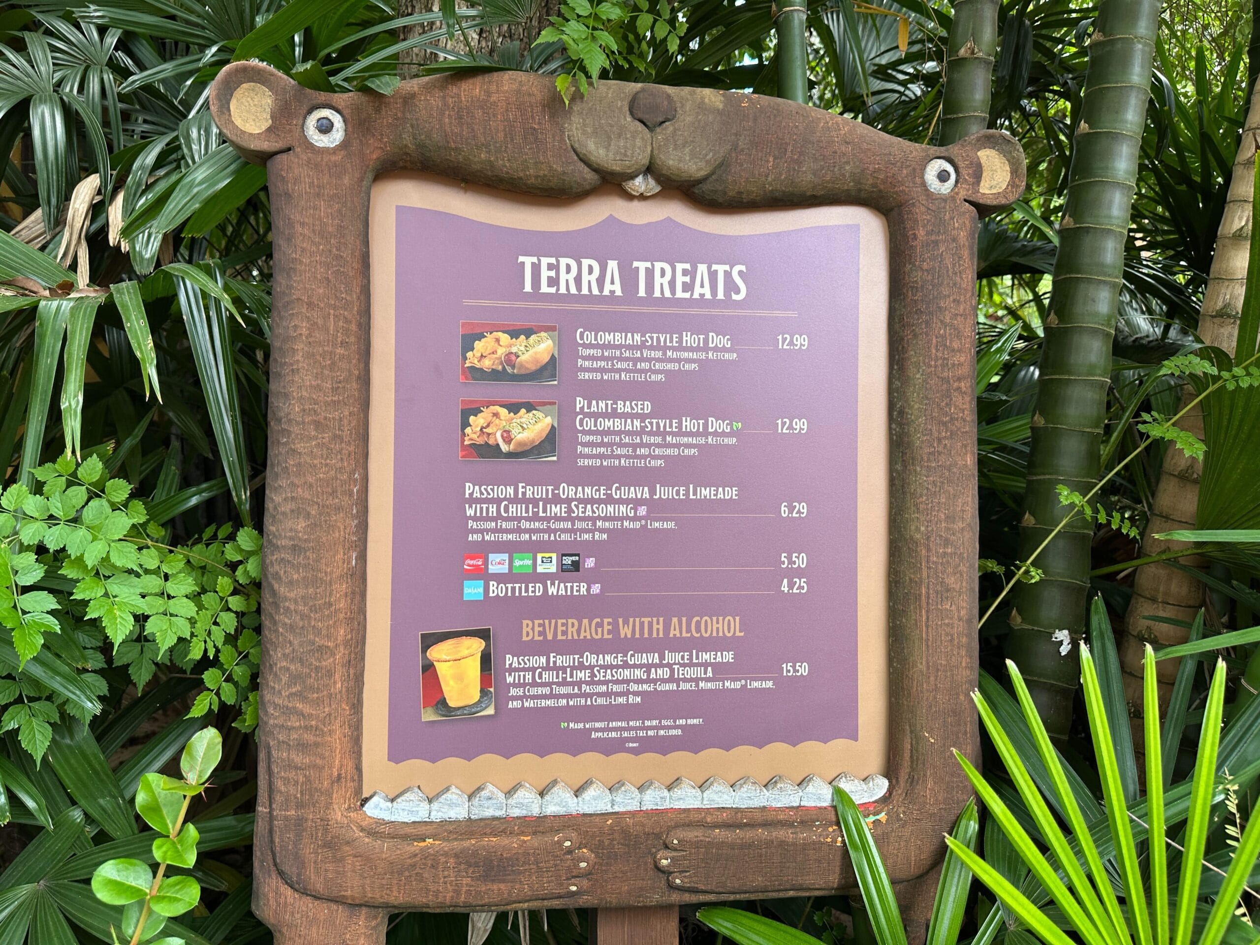 The Terra Treats menu board at Disney, framed by lush plants, features a Colombian-Style Hot Dog, POG Limeade, and bottled water.
