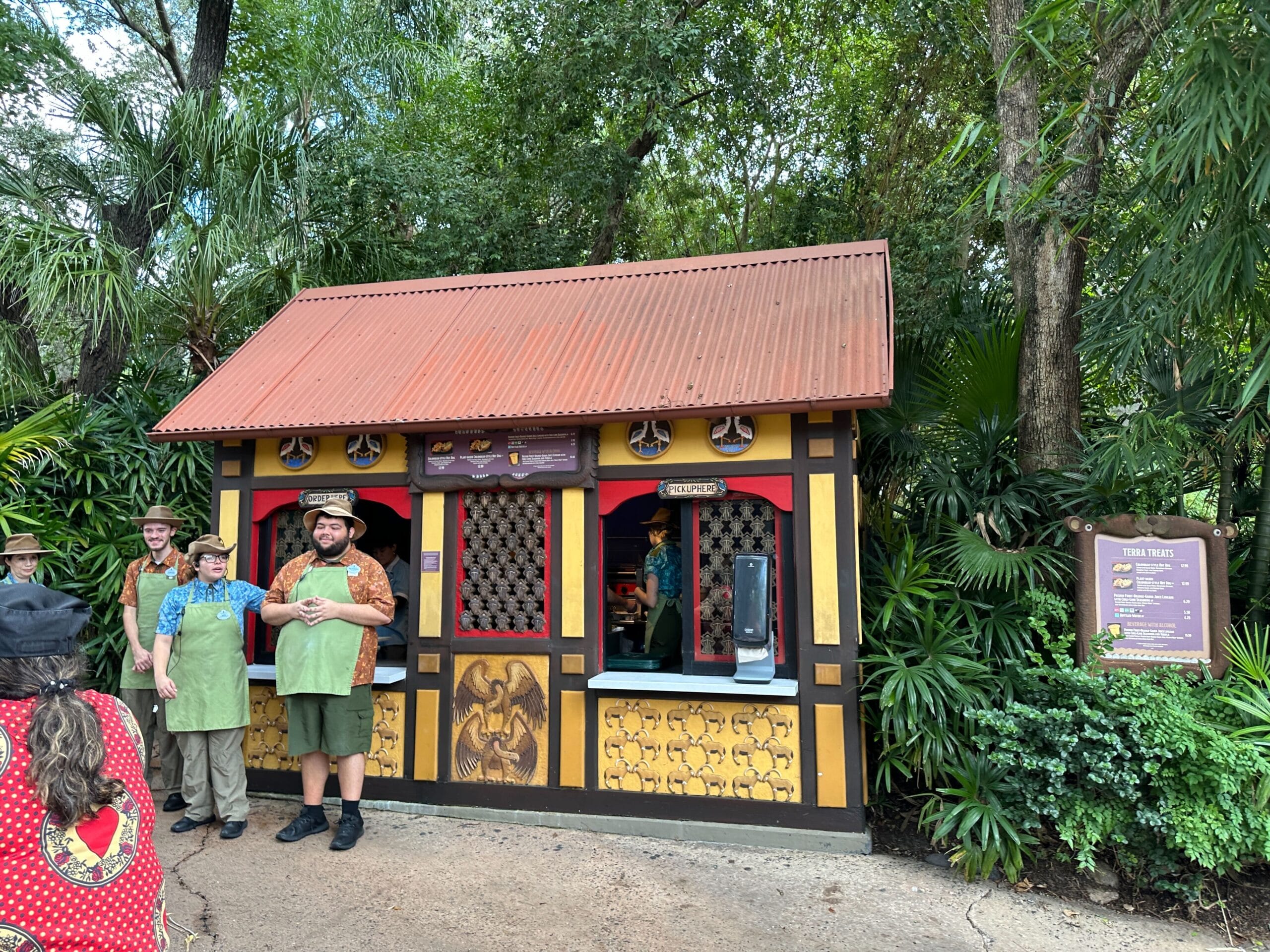 A small red-roofed food stand serves Colombian-Style Hot Dogs, nestled in greenery with a crowd—reminiscent of Disney park eateries.