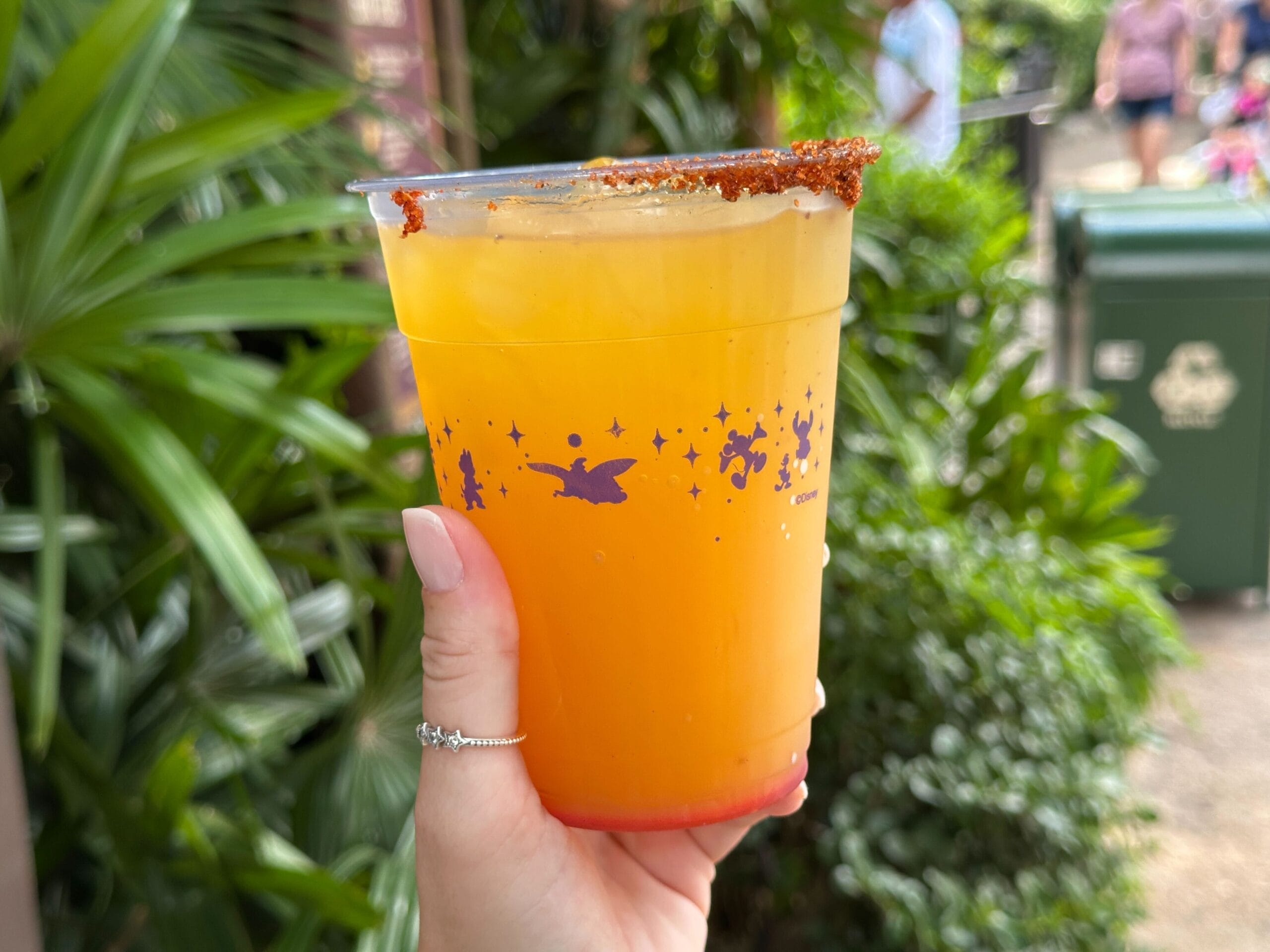A hand holds a POG Limeade in a plastic cup with a chili powder rim, outside by lush greenery