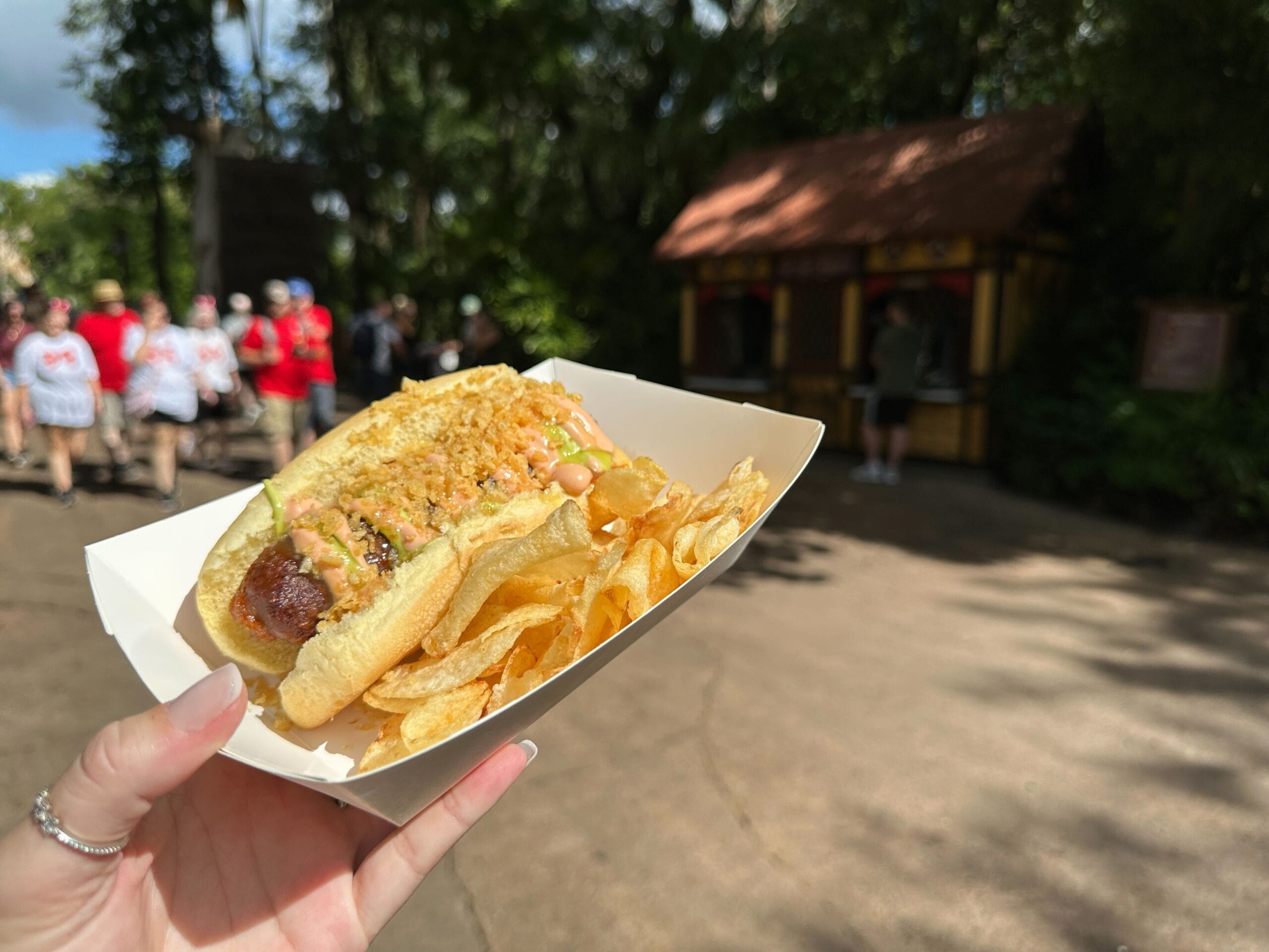 A hand holds a plant-based Colombian-Style Hot Dog with sauce and chips at Terra Treats in a lively, tree-filled Disney park setting.