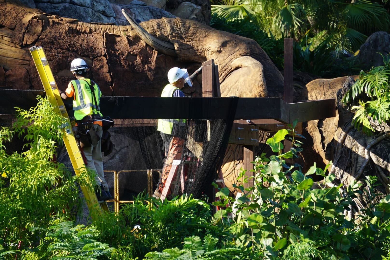Two construction workers in safety gear install signage beside artificial rocks for the Zootopia Better Zoogether Show at a Disney park.