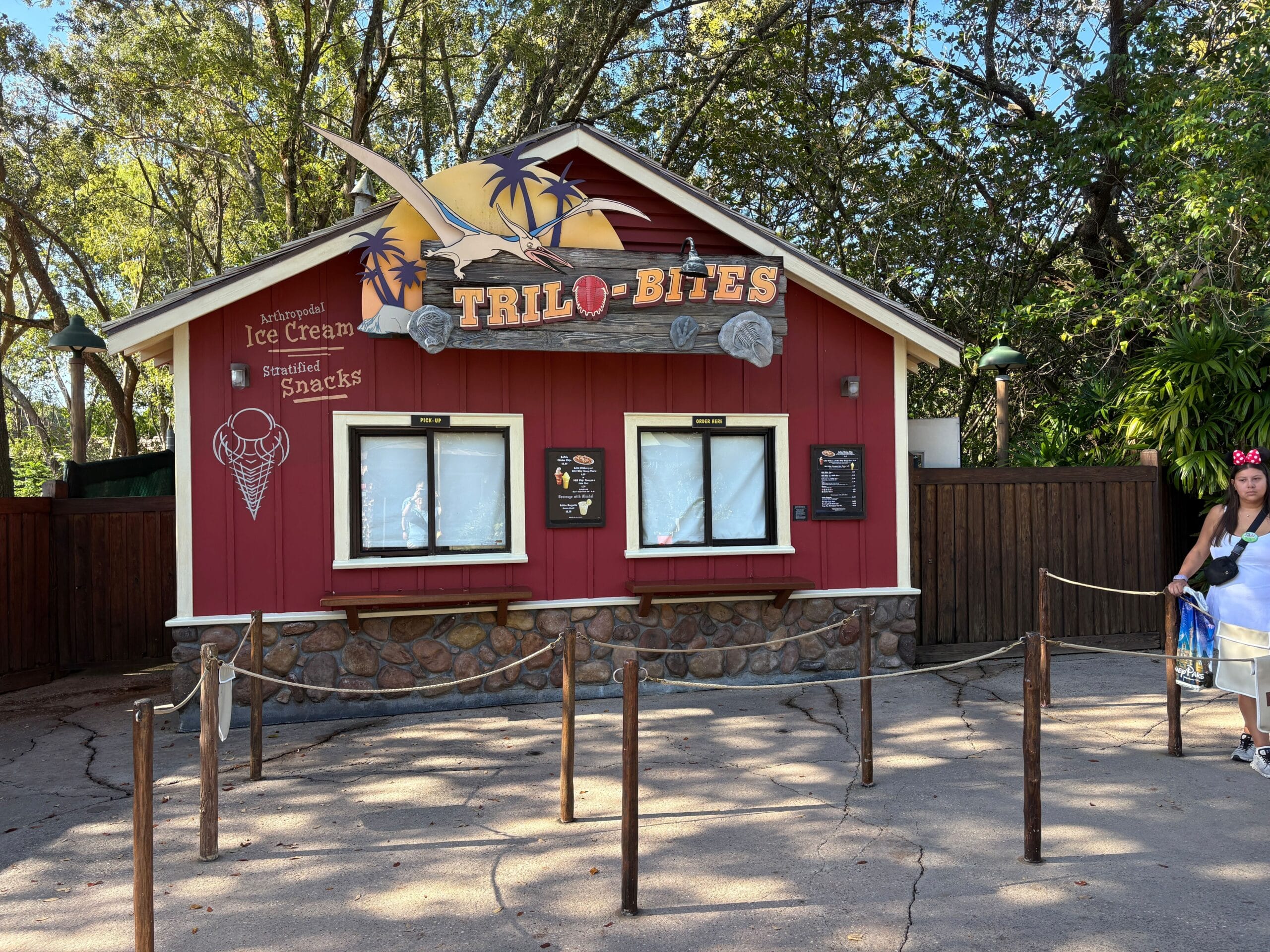 Trilo-Bites is a vibrant red snack stand with playful dinosaur decor, menus, and a rope queue in Animal Kingdom, not near any castle.
