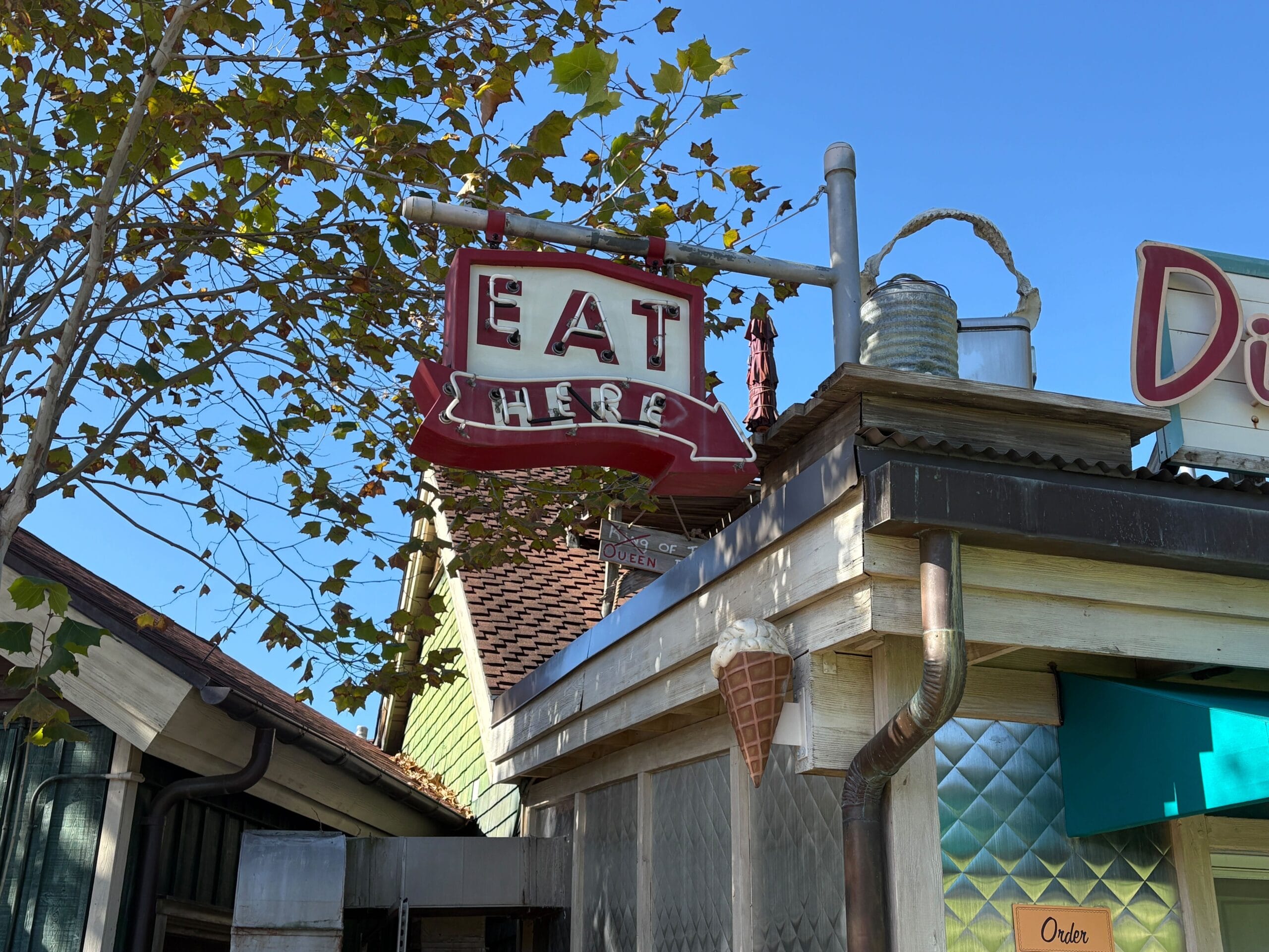 Red and white "EAT HERE" sign above Trilo-Bites diner in Disney's Animal Kingdom, with ice cream cone, tree, and blue sky overhead.