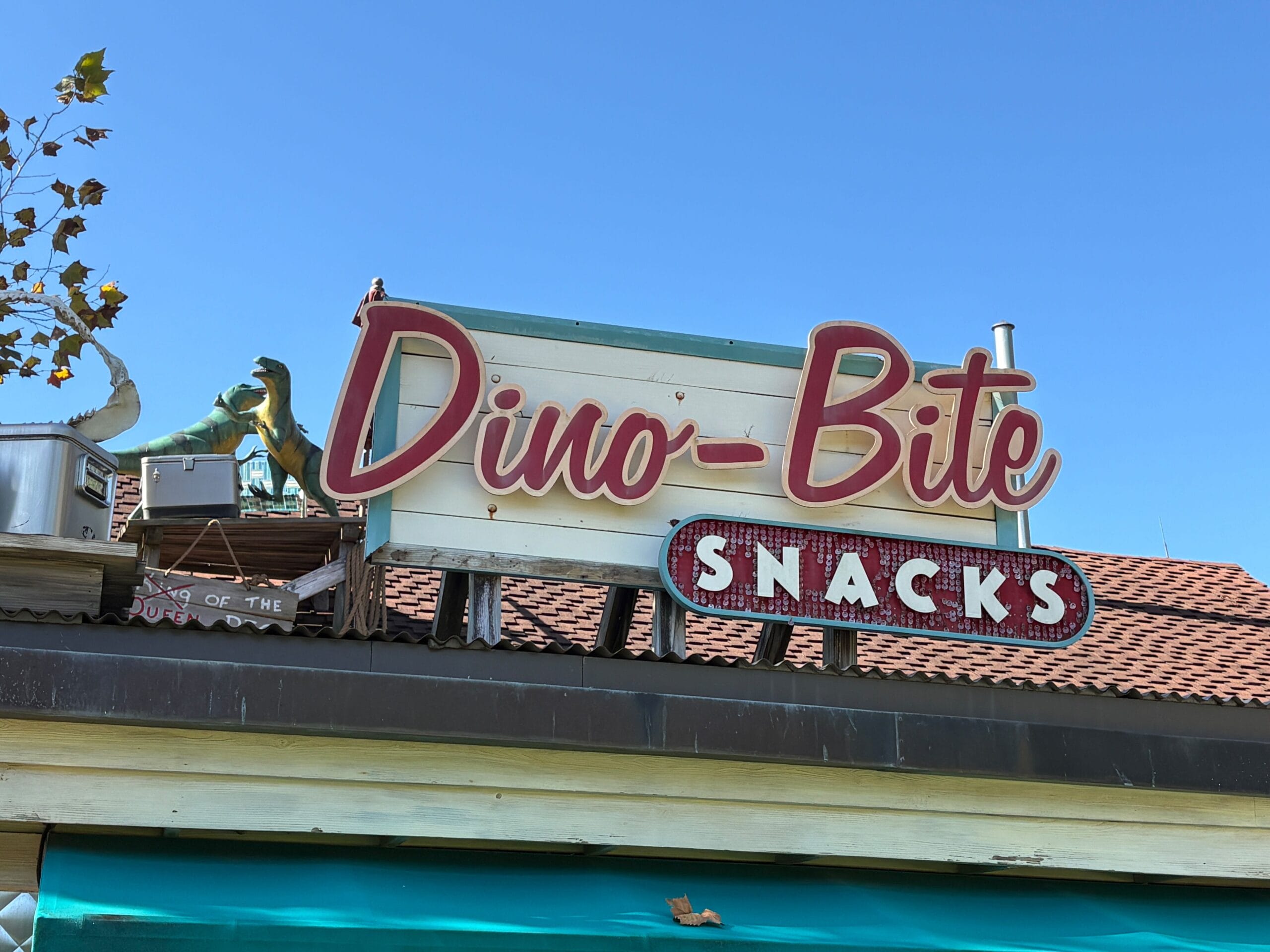 A vintage Dino-Bite Snacks sign with playful dinos on the roof at Disney's Animal Kingdom, set against a clear blue sky. No castle visible.