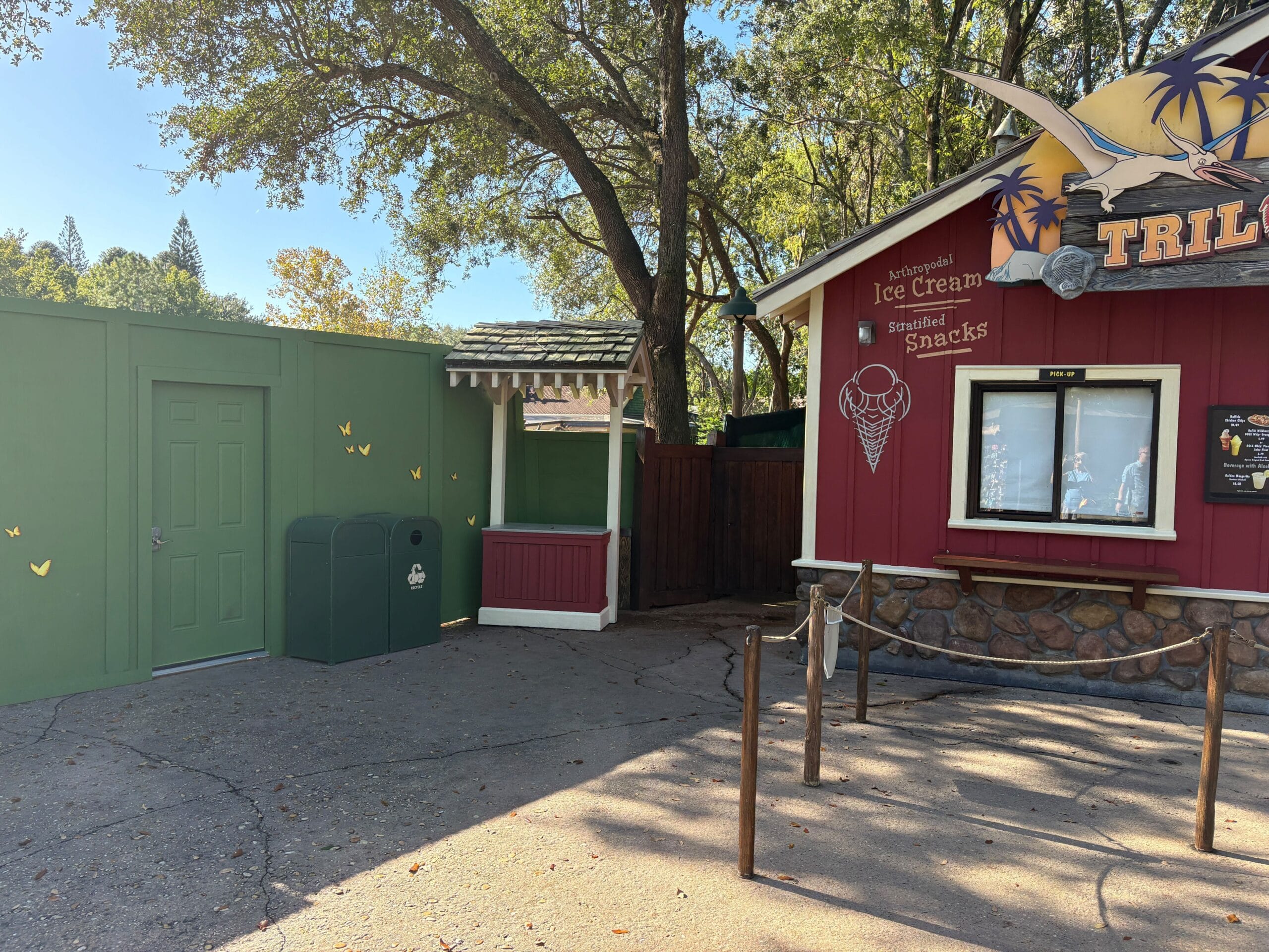 The closed Dino Bite Snacks stand and a green construction wall beneath trees in Disney's Animal Kingdom on a bright, sunny day.