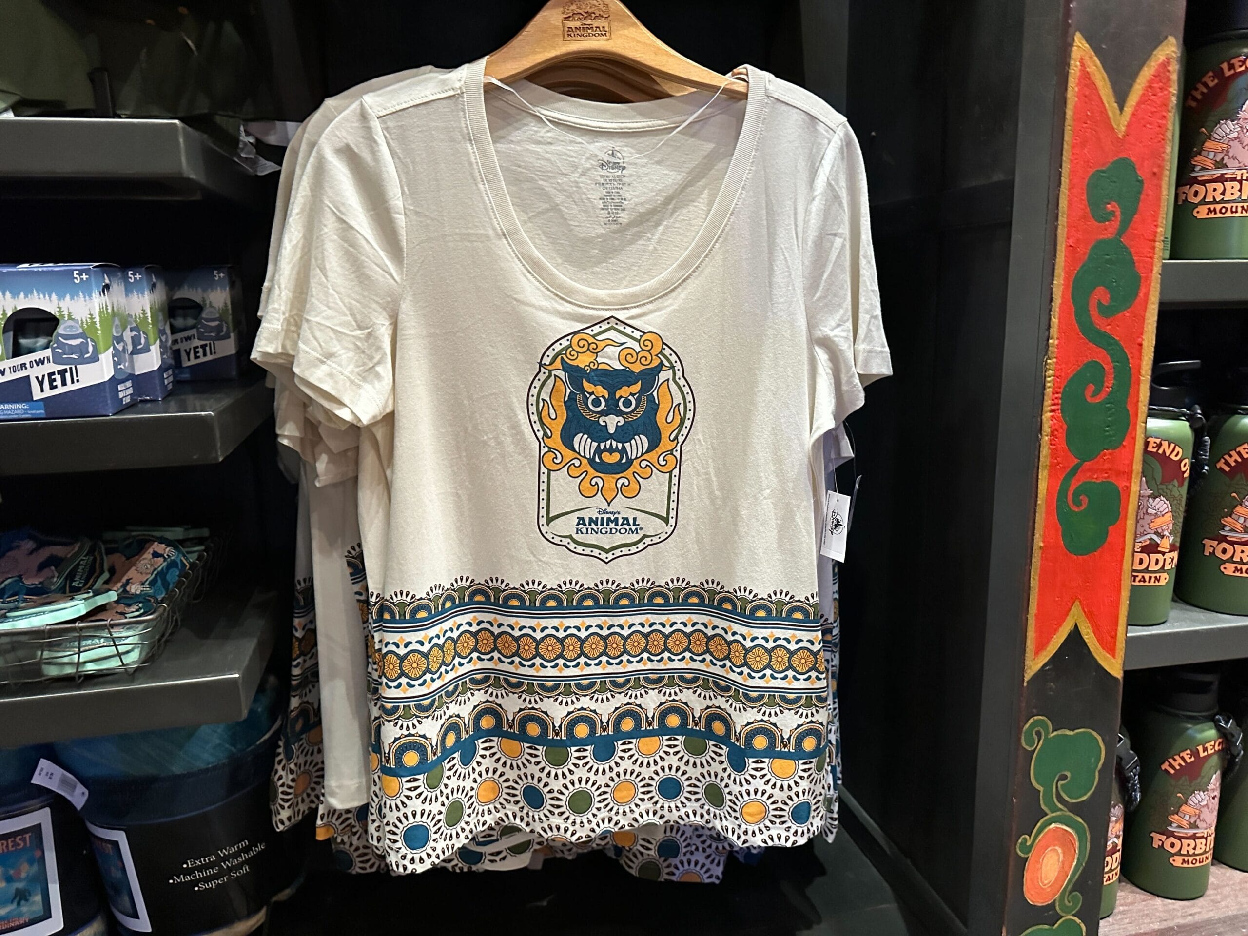 White t-shirt with a vibrant Surfer Yeti face and patterned trim, hanging on a Disney World rack near Cinderella Castle merchandise.