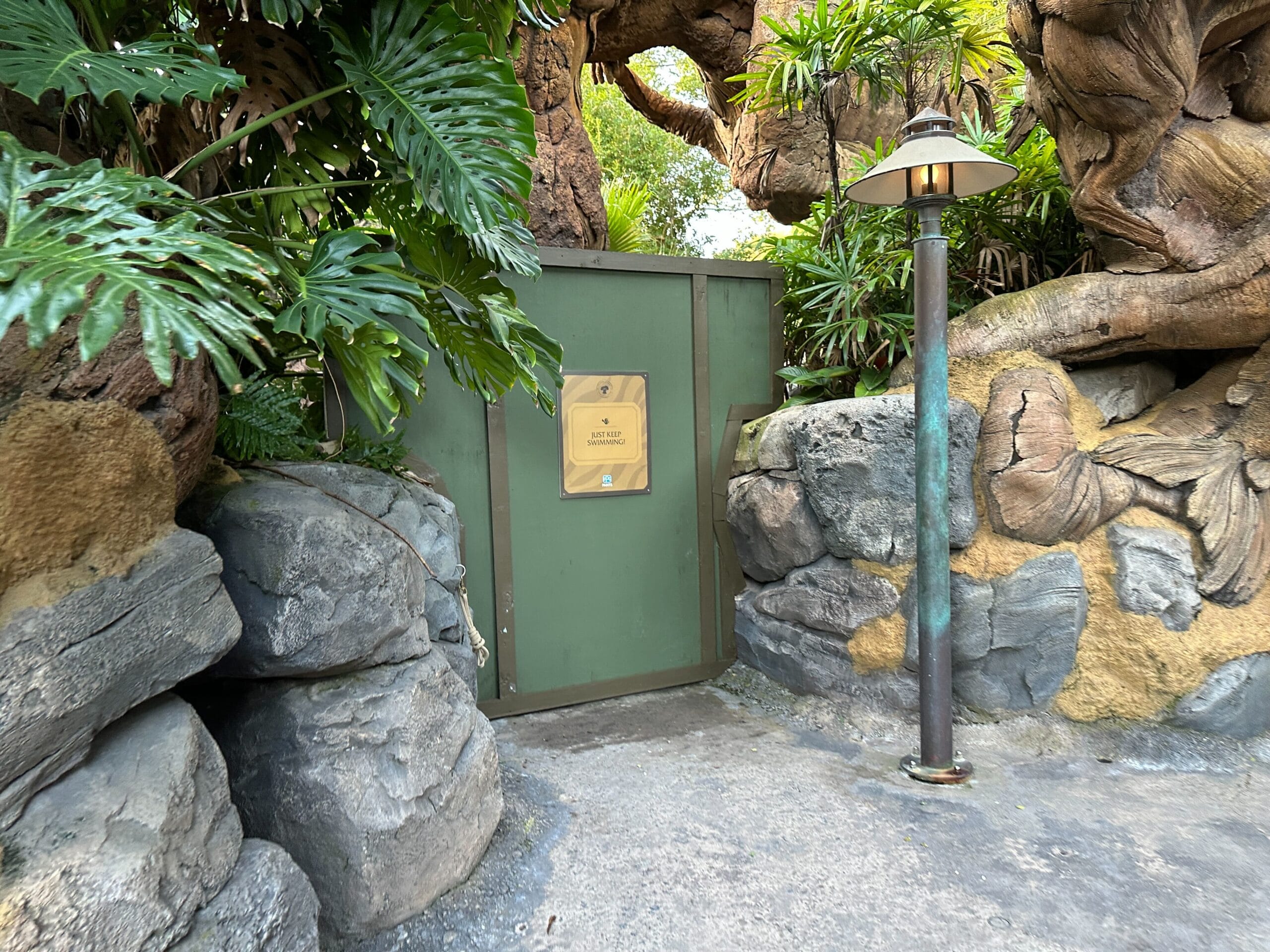 A green construction wall with a Show Marquee sign blocks a path between rocks and lush plants; a lamppost stands nearby.