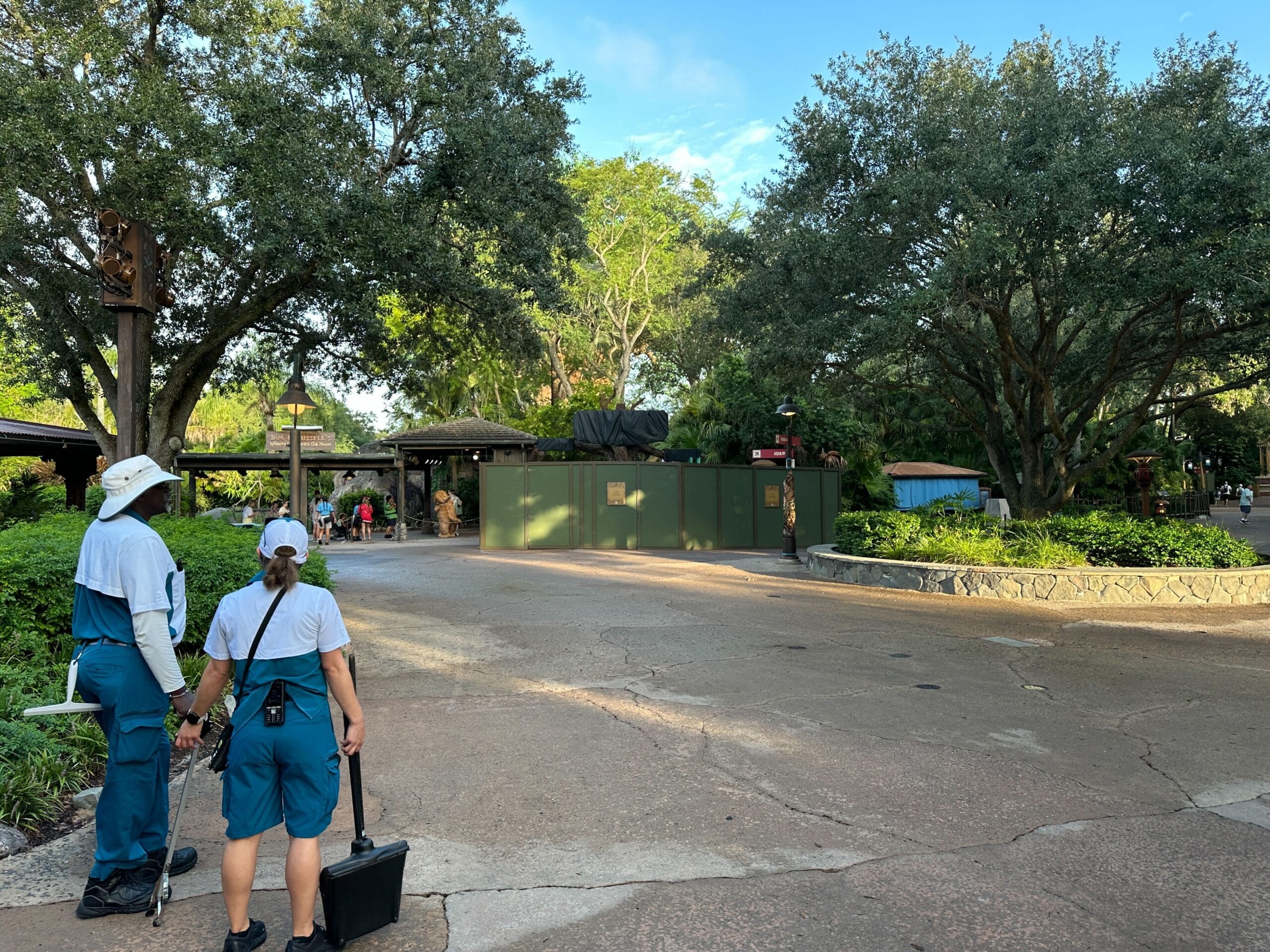 Two workers stand by a green construction wall in a tree-lined park at Animal Kingdom on a sunny day.