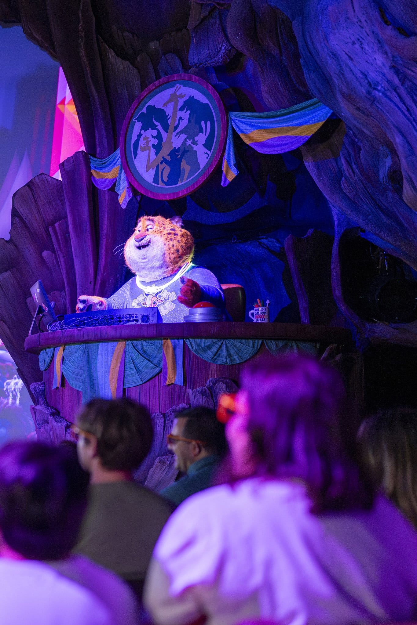 A Zootopia tiger DJ entertains a lively Disney Park crowd, performing beneath an entrance sign, no iconic castle in view.