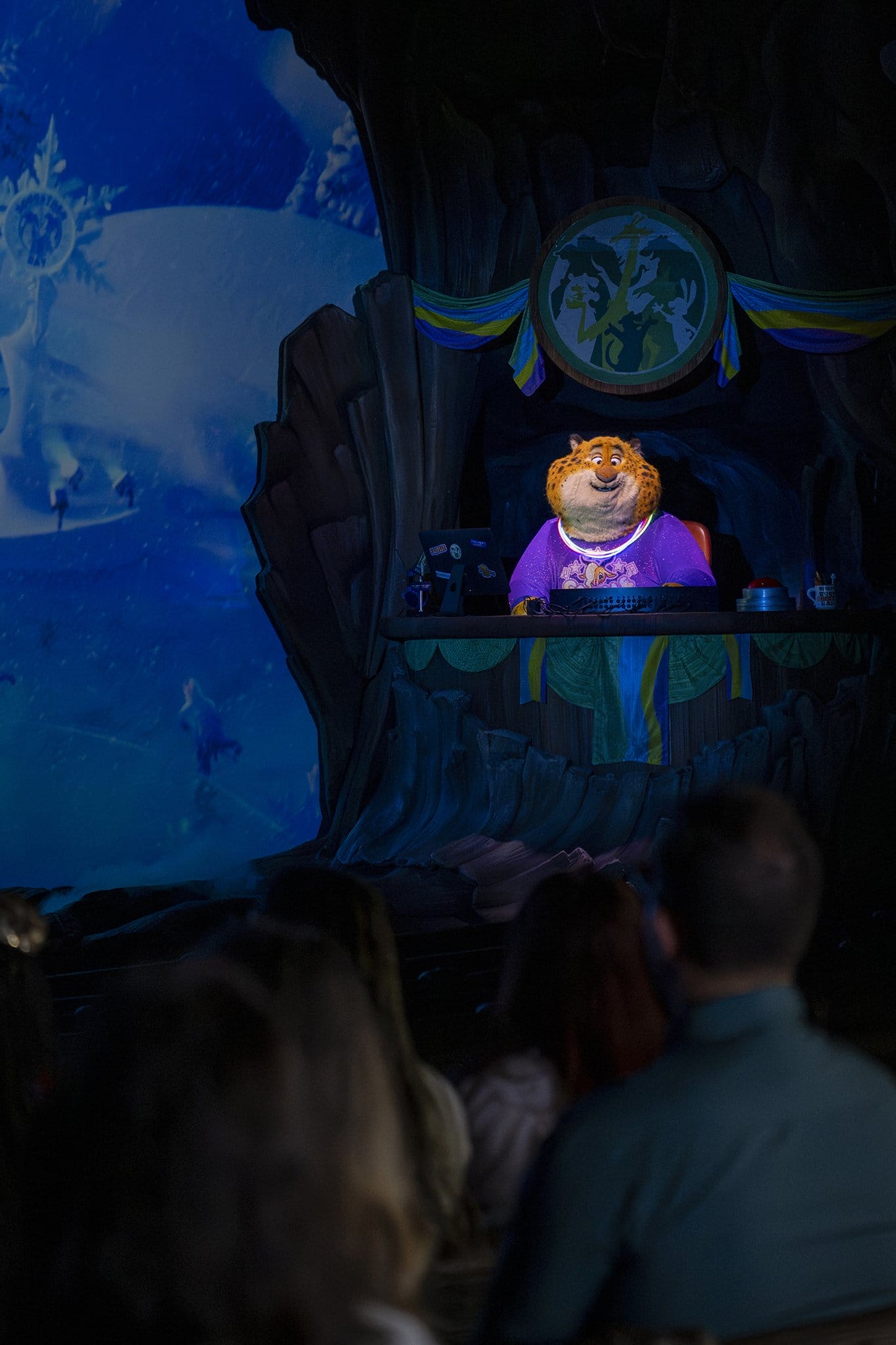 A Zootopia character entertains guests on a Disney Parks stage with winter decorations and a park entrance sign in the background.