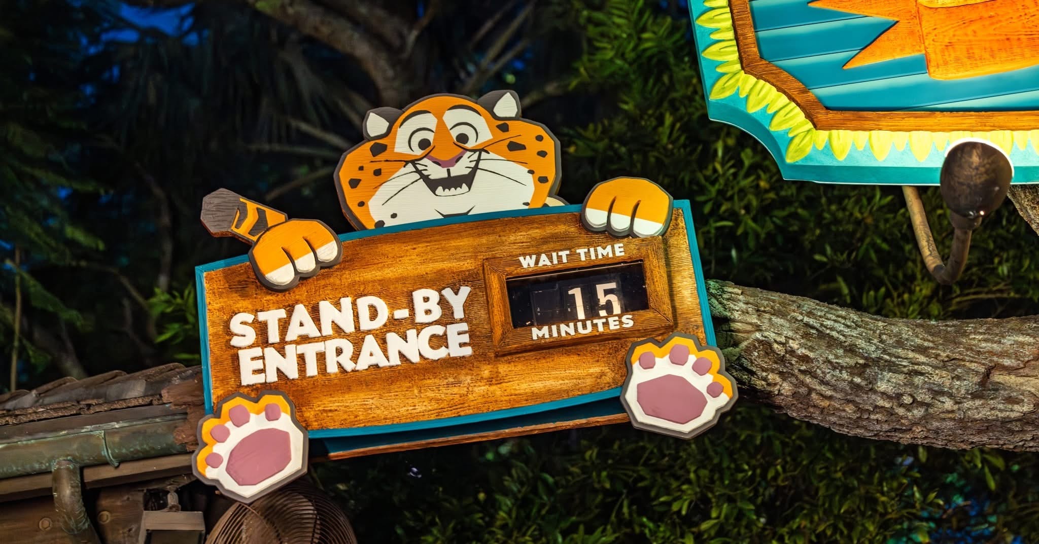 Cartoon tiger at a Disney park holding a "Stand-By Entrance" 15-minute wait sign, surrounded by themed trees and lush foliage.