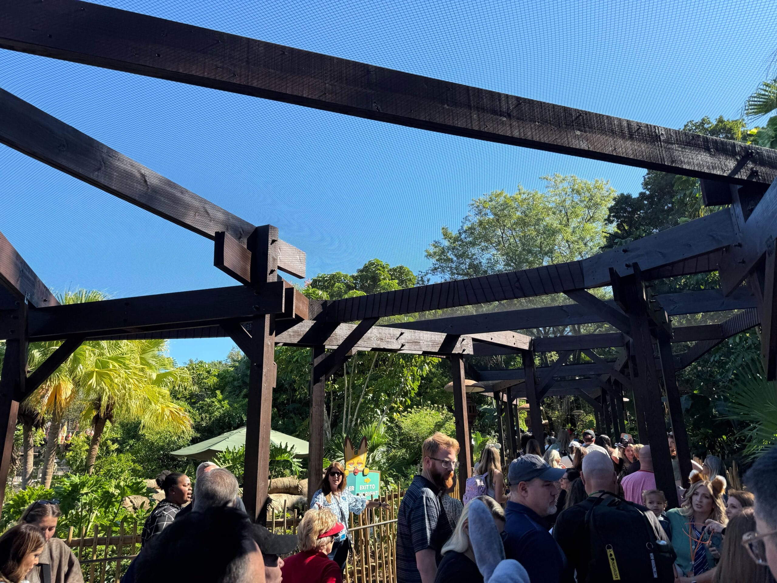 Guests gather beneath rustic wooden beams in the sun at Disney’s Animal Kingdom, nestled among lush, tropical foliage.