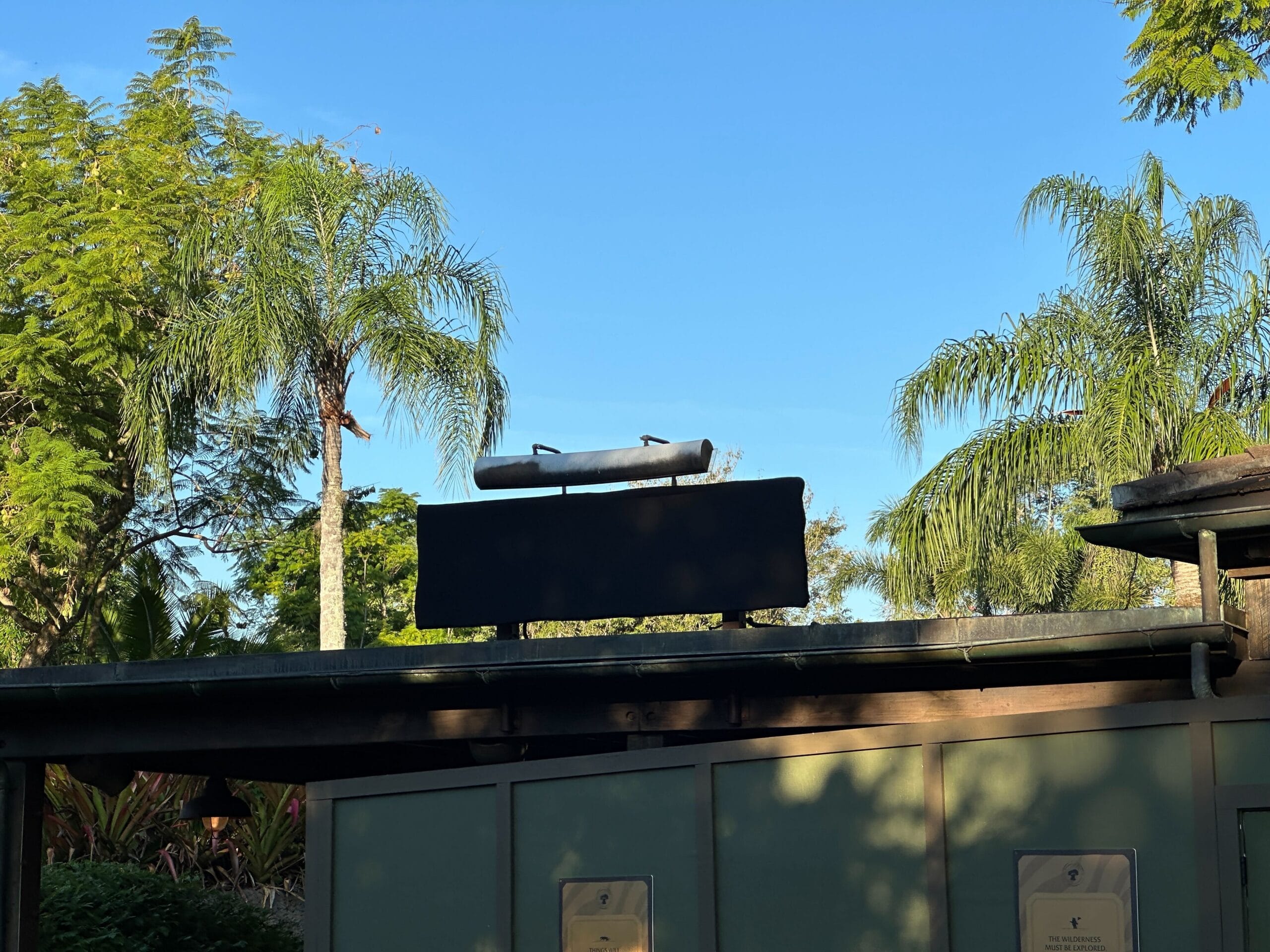 A blank black sign on a rooftop evokes Zootopia vibes, framed by palm trees and a sky reminiscent of sunny Disney park days.