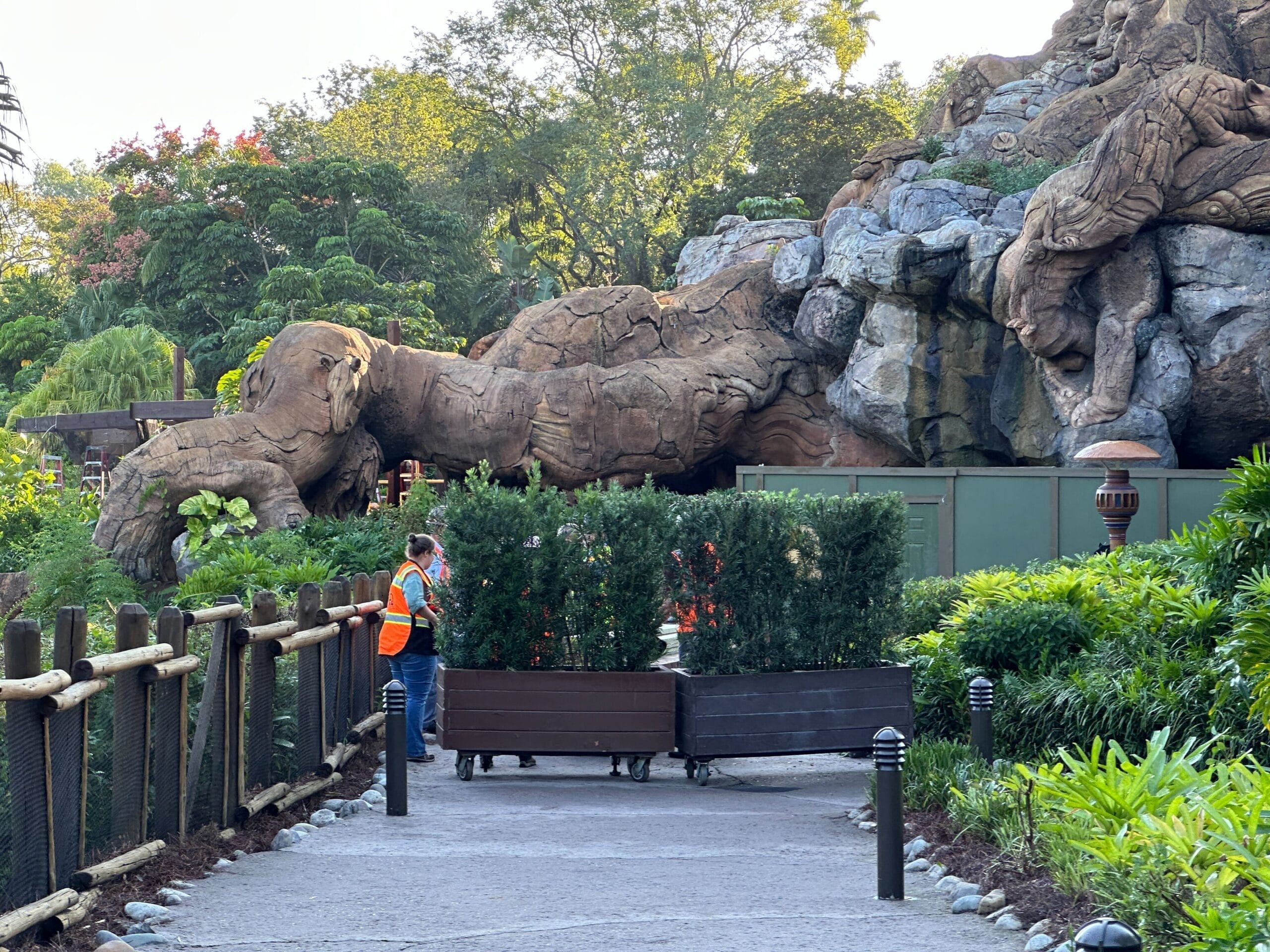 A worker in an orange vest stands by planters blocking a path to Epic Universe, in a park with greenery reminiscent of Disney parks.