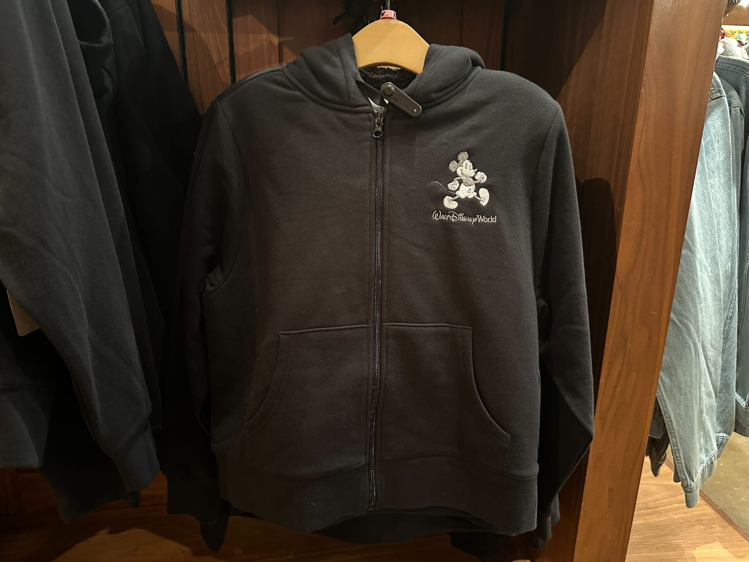 Black Sherpa hoodie featuring a small Mickey Mouse logo and "Walt Disney World" text, displayed on a wooden rack at a Disney park shop.