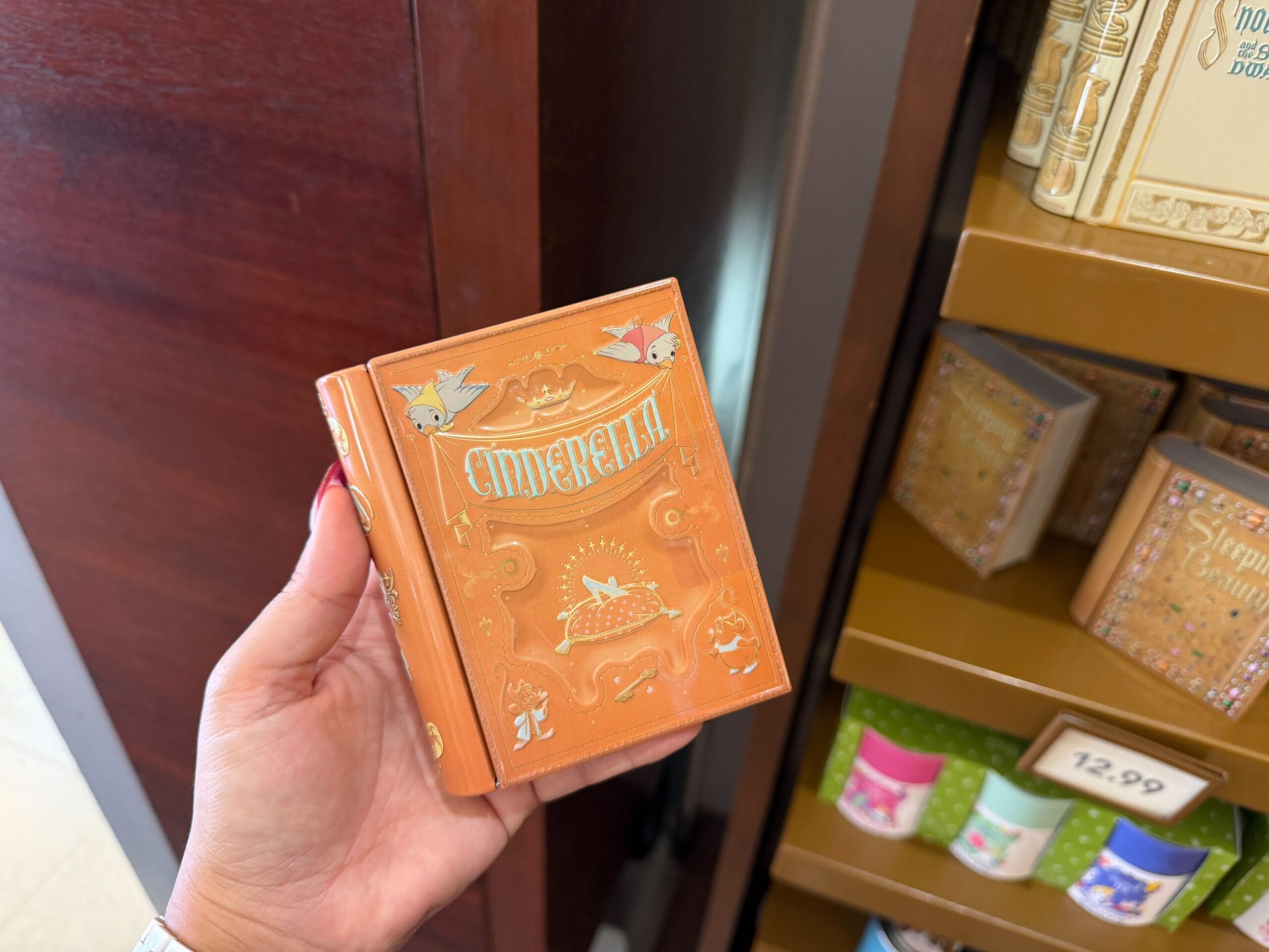 A hand displays a small, ornate orange "Cinderella" storybook tin in front of a Disney park store shelf stocked with souvenirs.