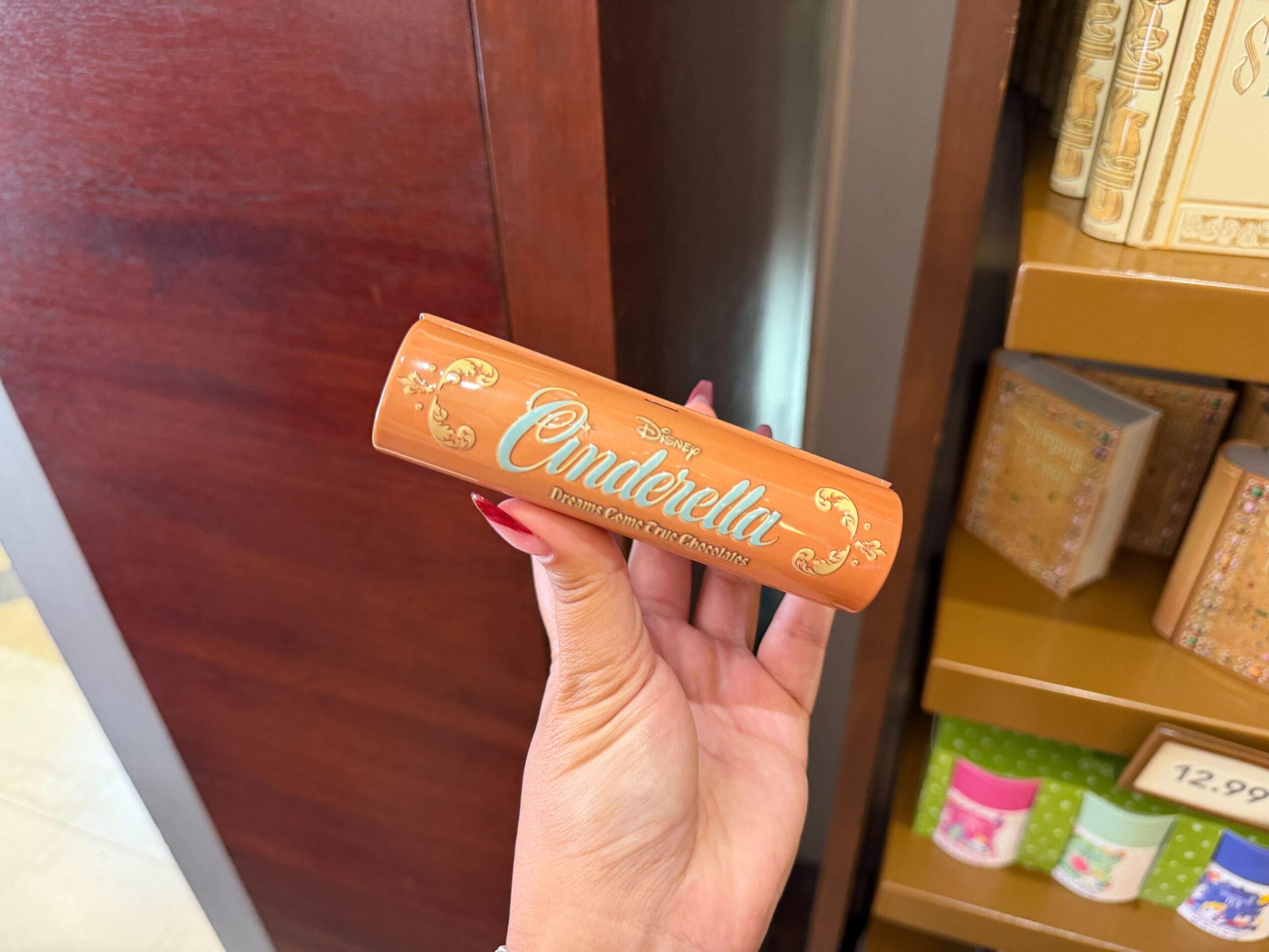 A hand with red nails holds a Cinderella Story Book Keepsake Tin compact inside a Disney World shop, near Cinderella Castle.