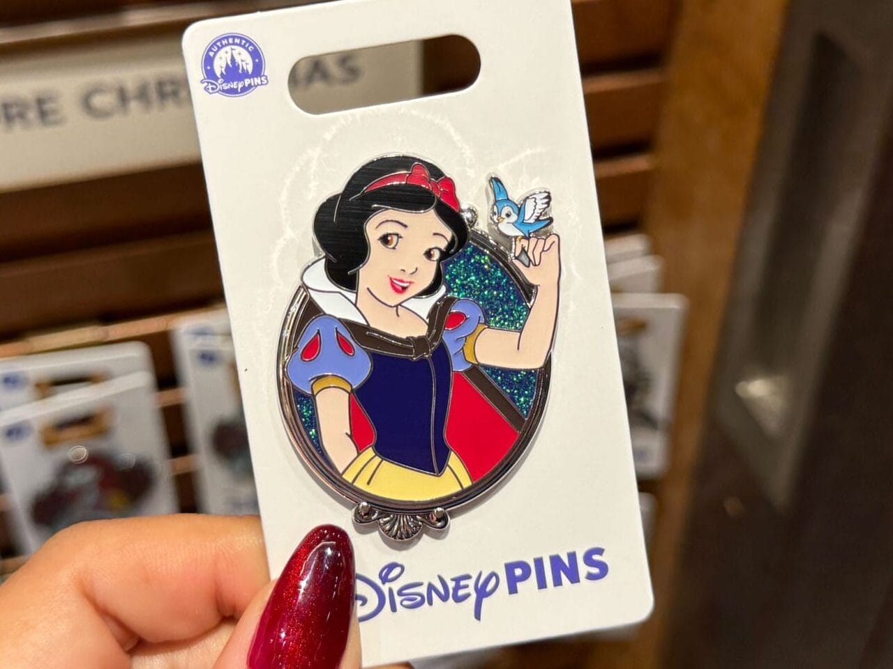 A hand displays a Snow White Princess pin in a Disney park shop, other character pins visible behind, evoking classic park merch.