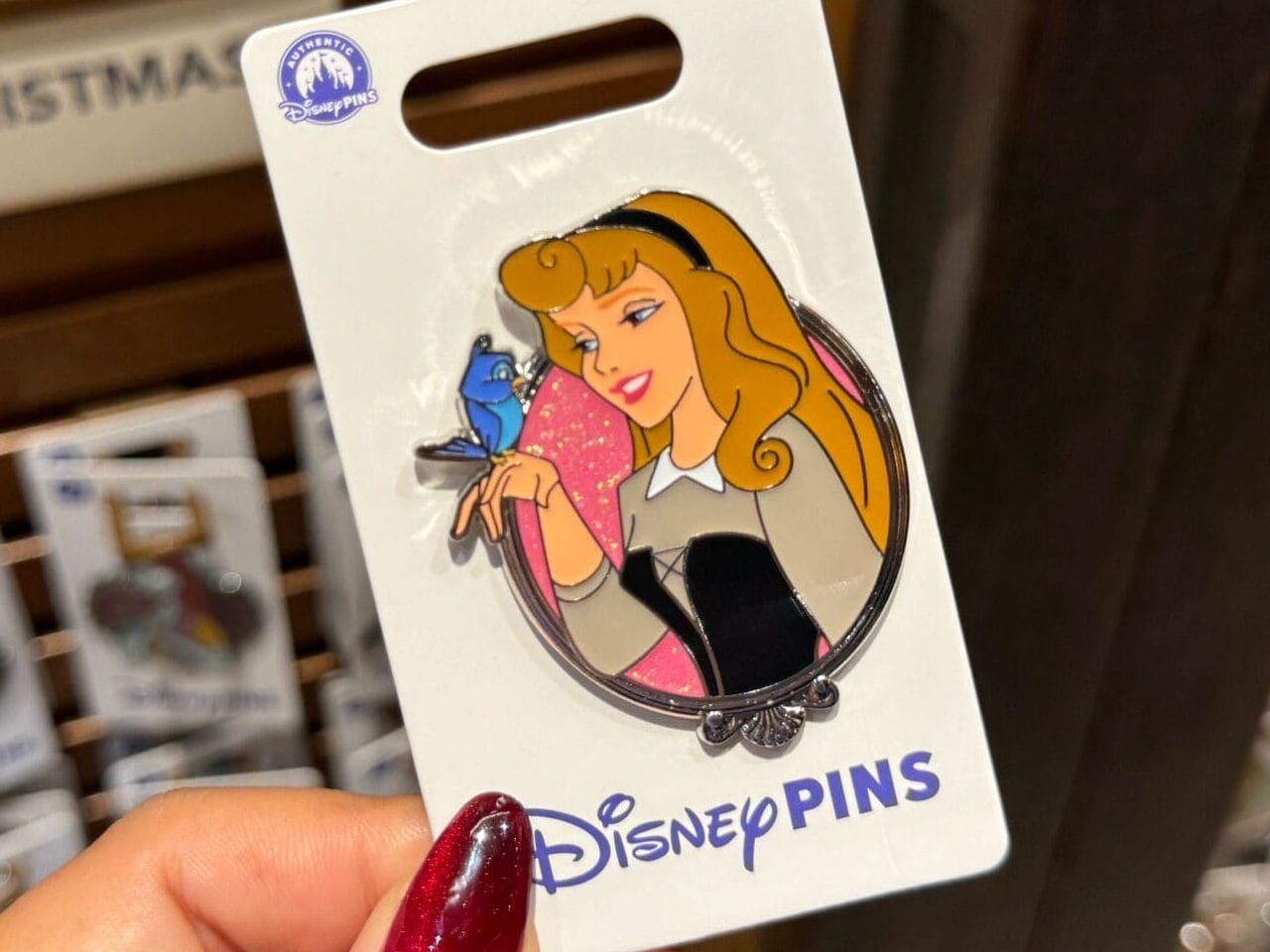 A hand holds a Disney Princess Aurora pin with a bluebird, surrounded by park-exclusive pins at an international Disneyland store.