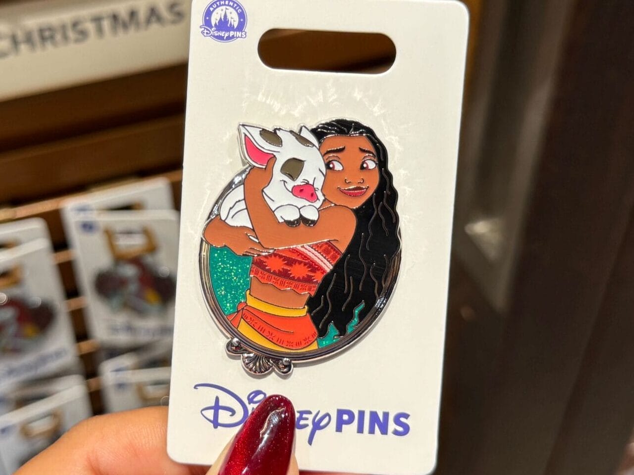 A hand displays a Moana and Pua princess pin among other Disney pins at a park shop—no castle visible for comparison.