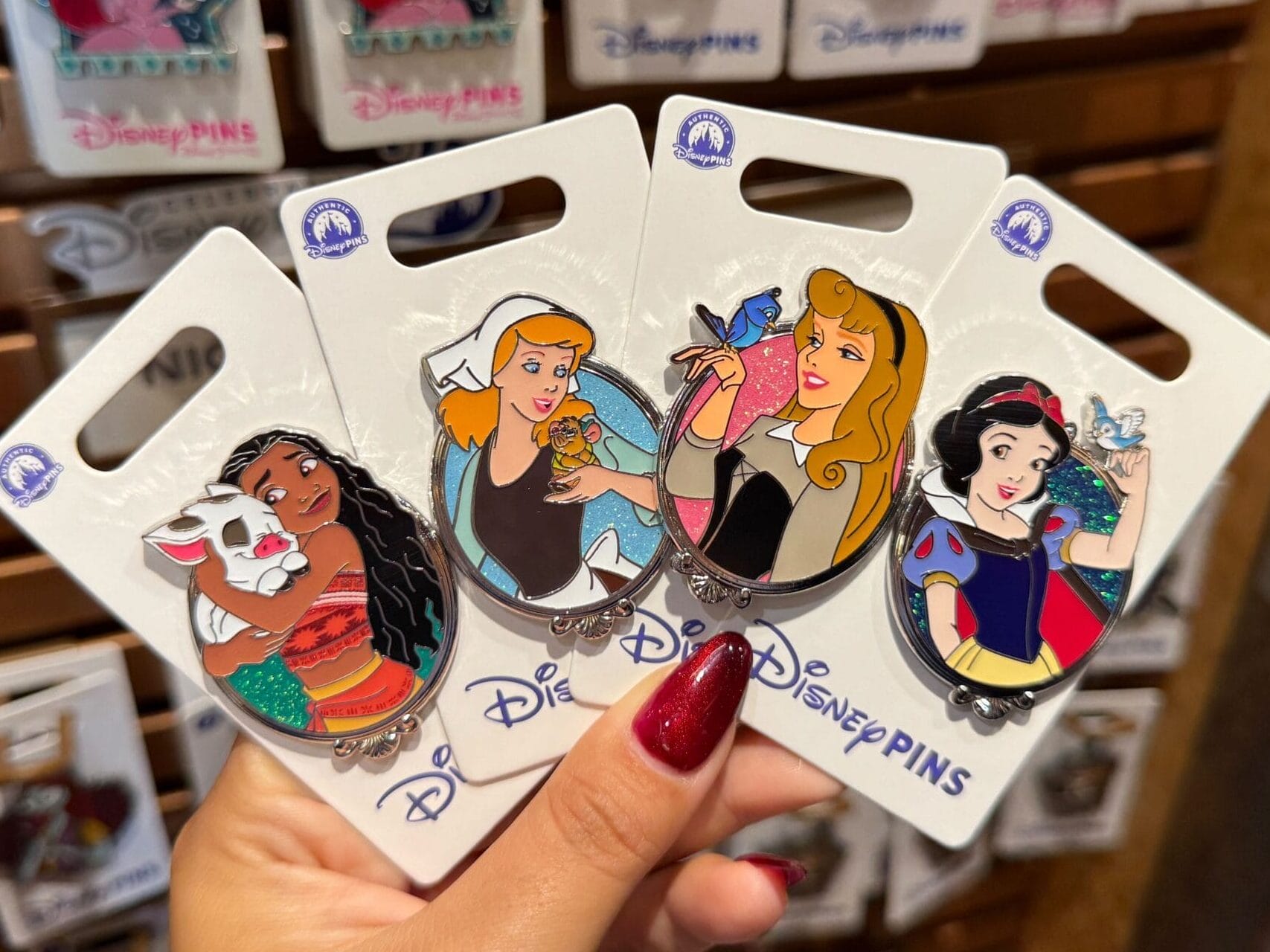 Inside a Disney Park shop, a hand displays four pins of Moana, Rapunzel, Aurora, and Snow White amid themed park decor.