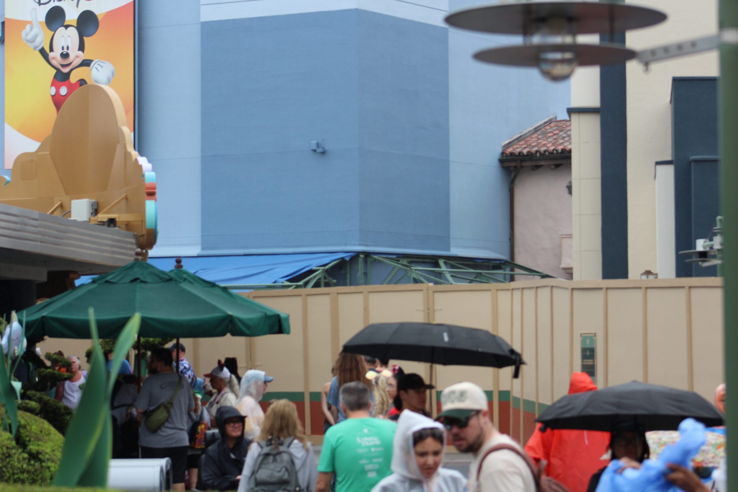 Guests with umbrellas gather by a demolition wall in Animation Courtyard at Hollywood Studios, Mickey Mouse sign in view.
