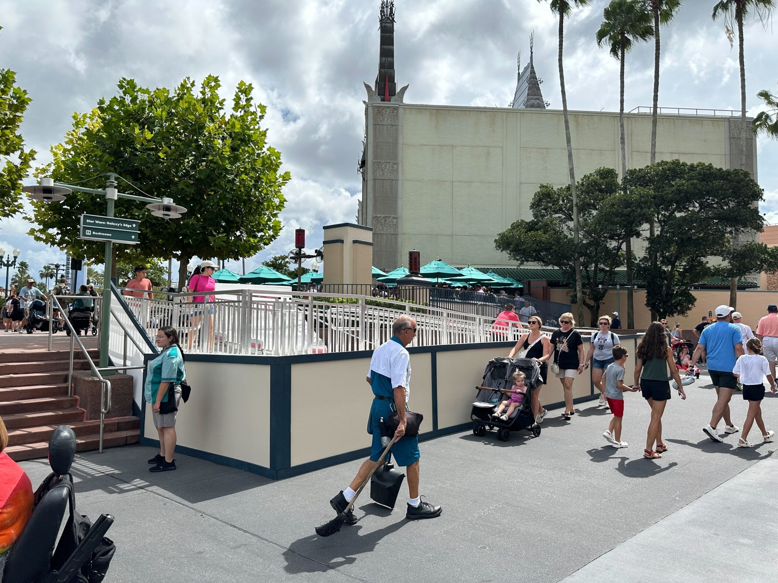 Guests stroll past construction walls and green umbrellas under a cloudy sky at Disney's Hollywood Studios; no castle in view.