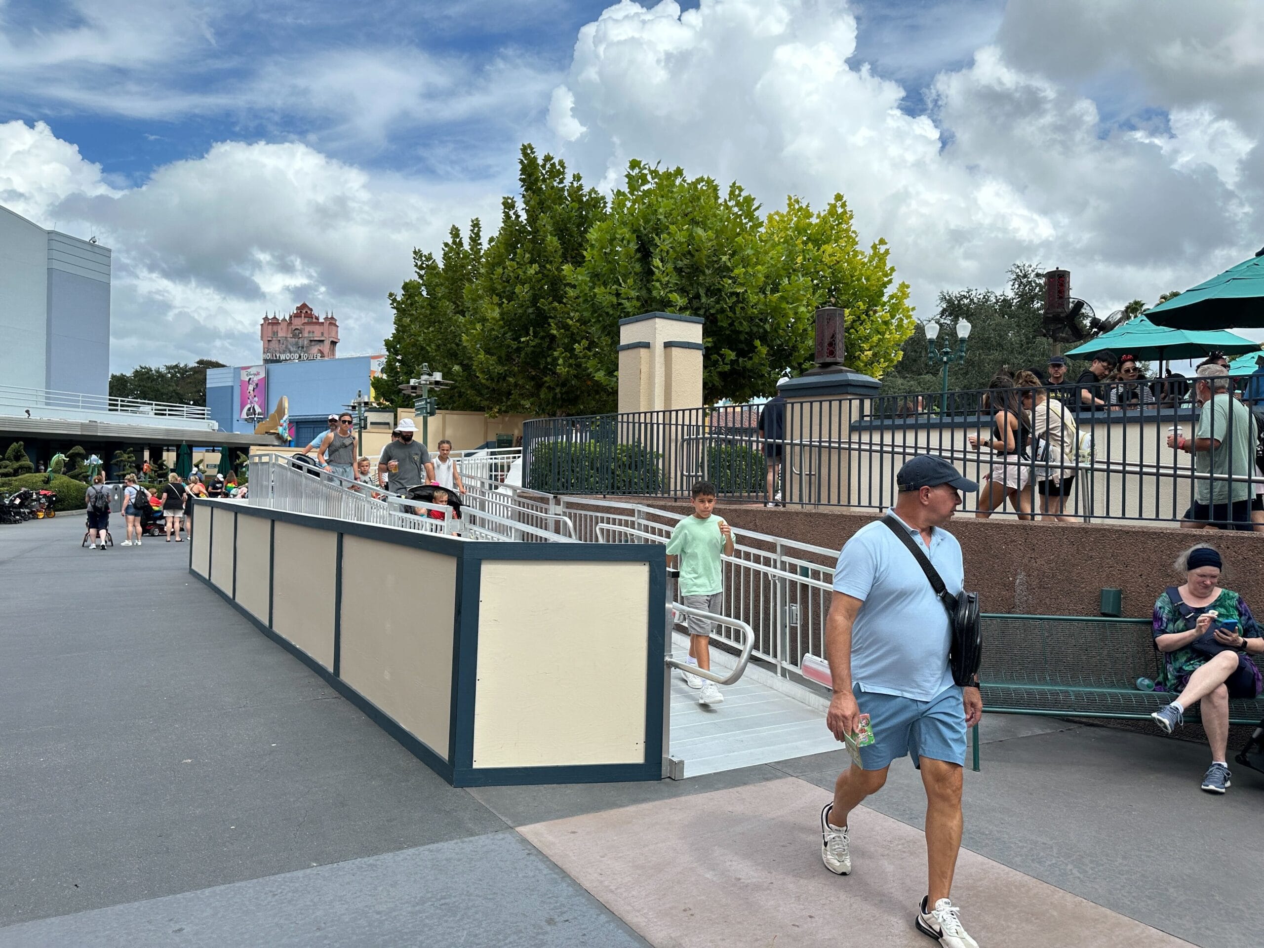 A man in a blue shirt walks past construction walls by a wheelchair ramp at Animation Courtyard, Disney’s Hollywood Studios. Cloudy sky.