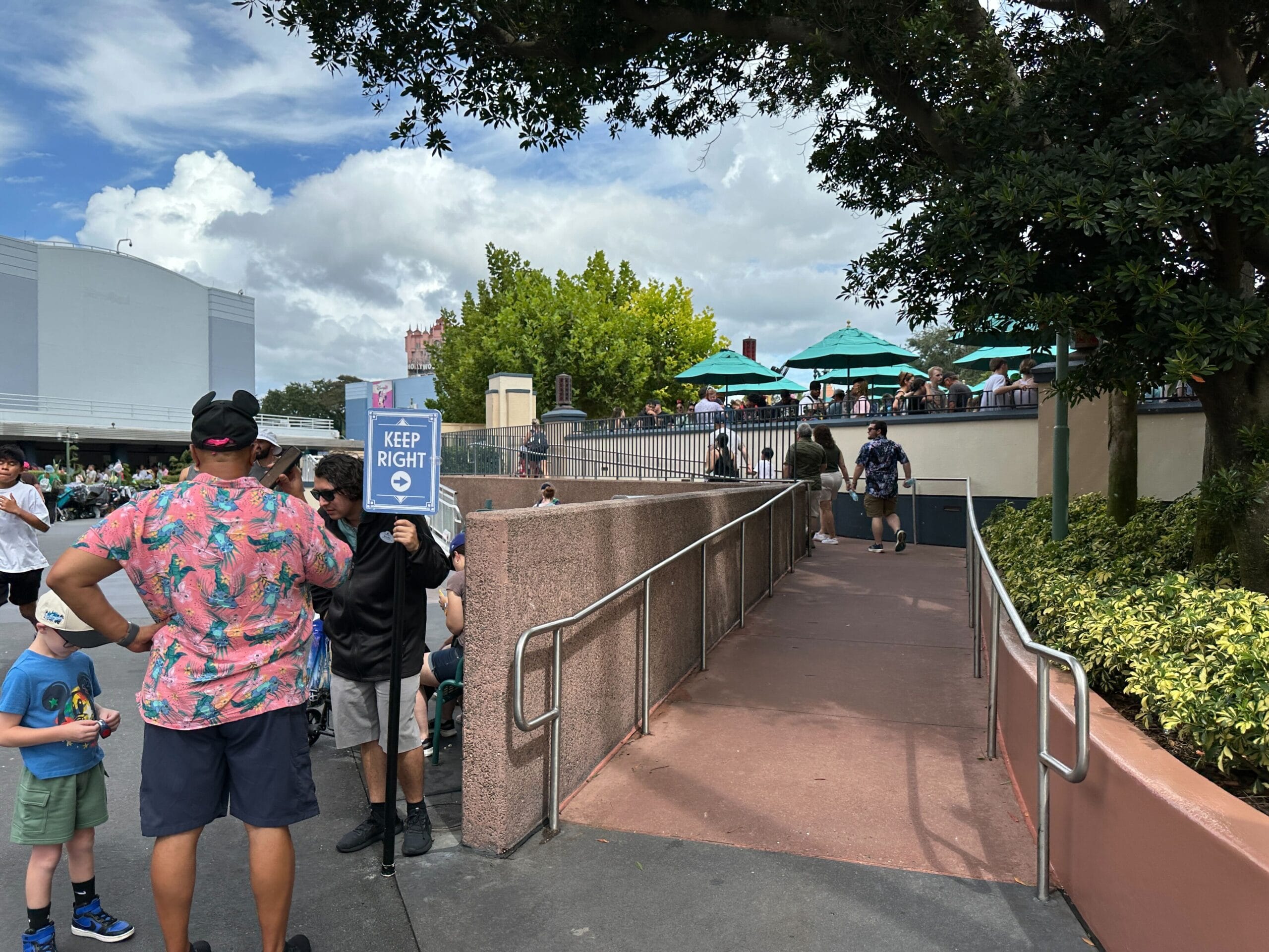 A person holds a "Keep Right" sign by construction walls in Animation Courtyard, guests relax under umbrellas like at Disney parks.
