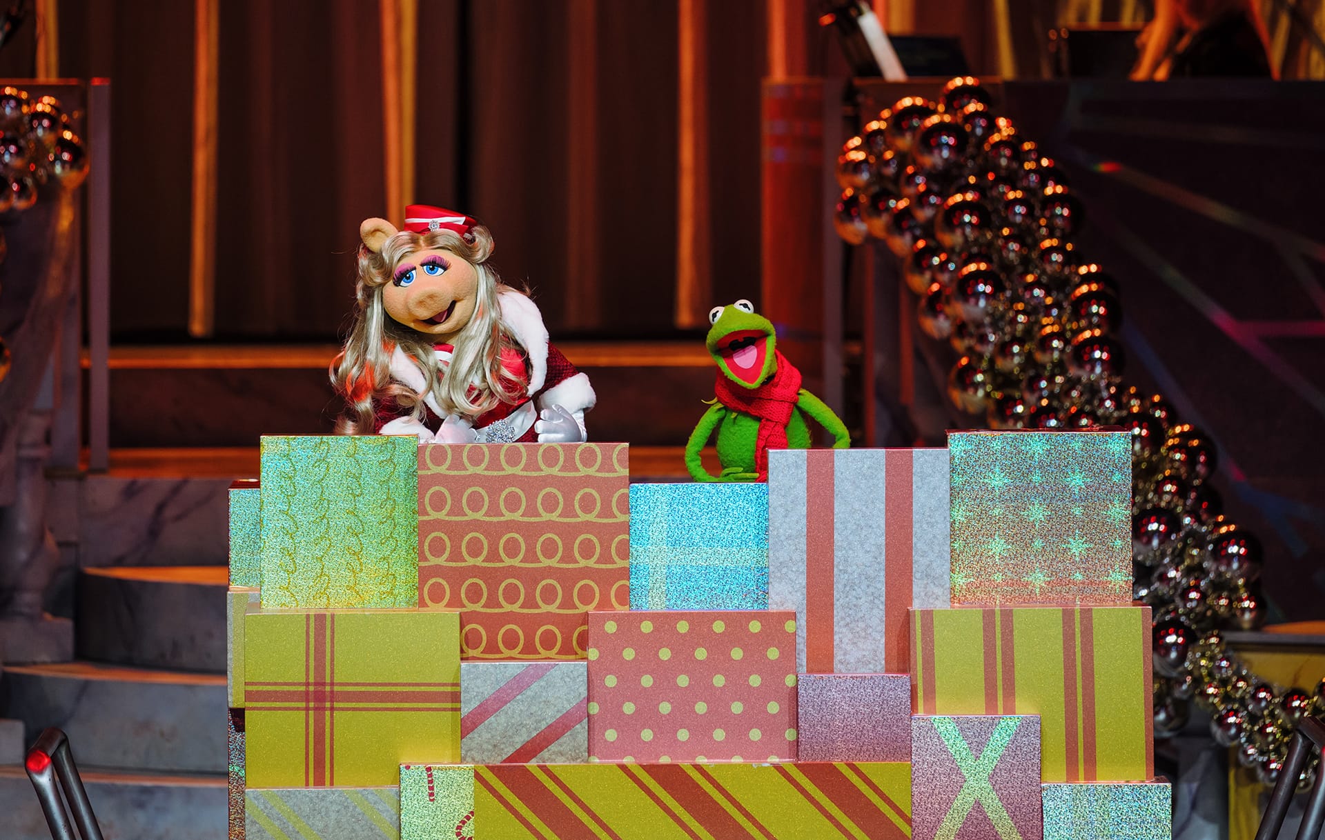 Miss Piggy and Kermit the Frog pose by vibrant presents on a sparkling Jollywood Nights 2025 stage at a Disney park event.