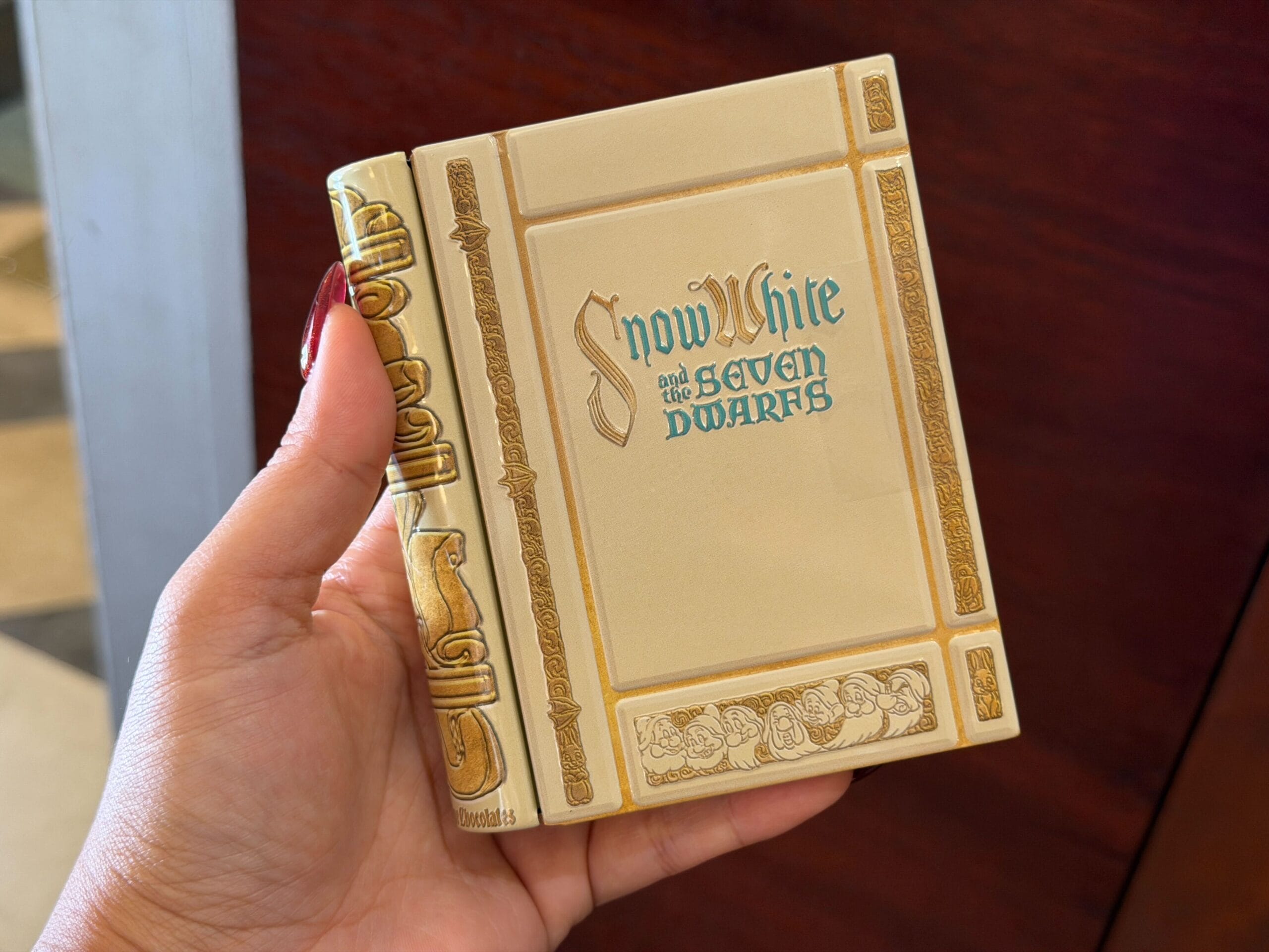 A hand holds a Disney Parks souvenir tin styled like a storybook, titled "Snow White and the Seven Dwarfs," with ornate gold borders.