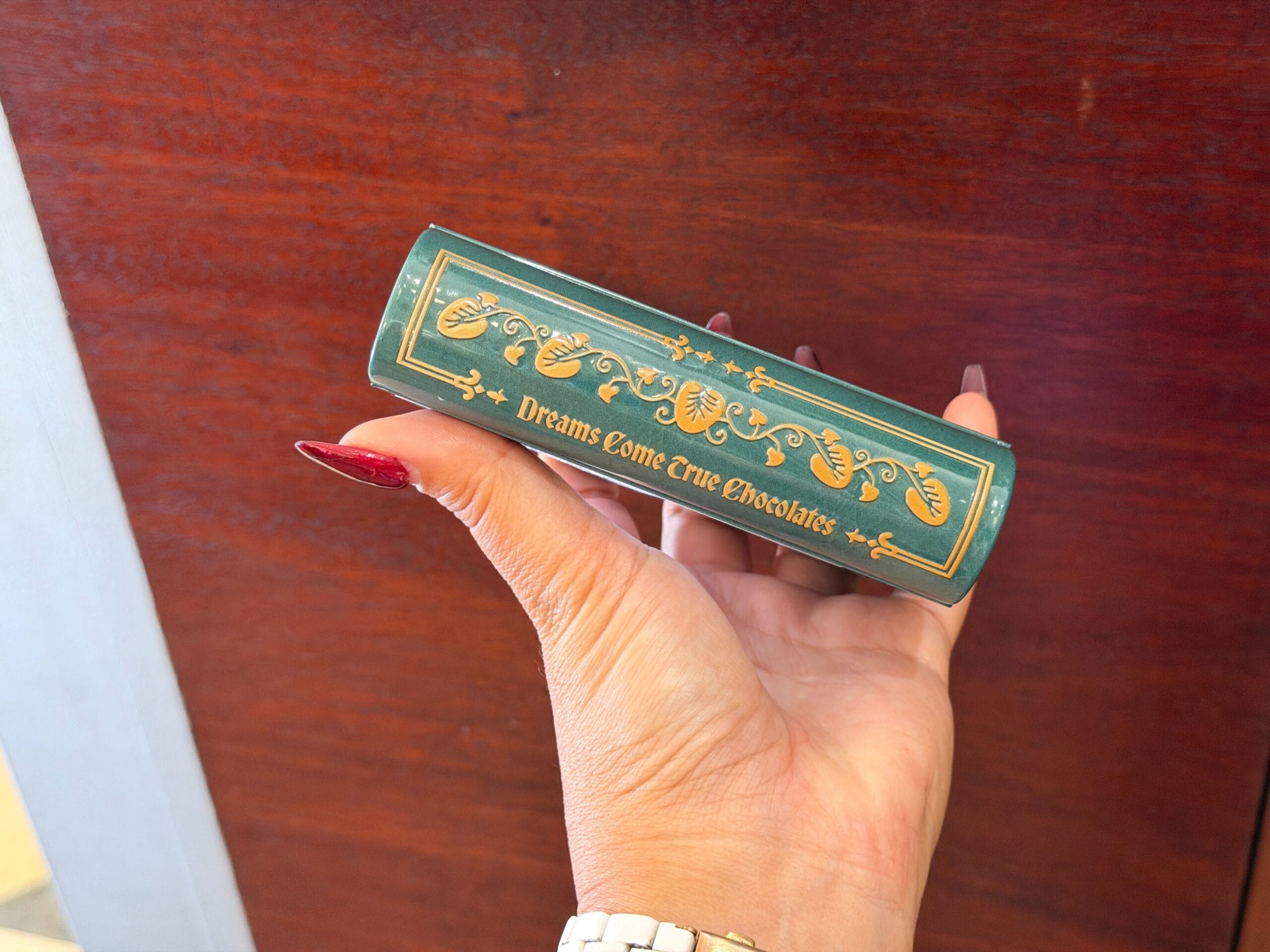 A hand with red nails holds a storybook tin of Dreams Come True Chocolates, featuring gold designs reminiscent of Disney Park souvenirs.