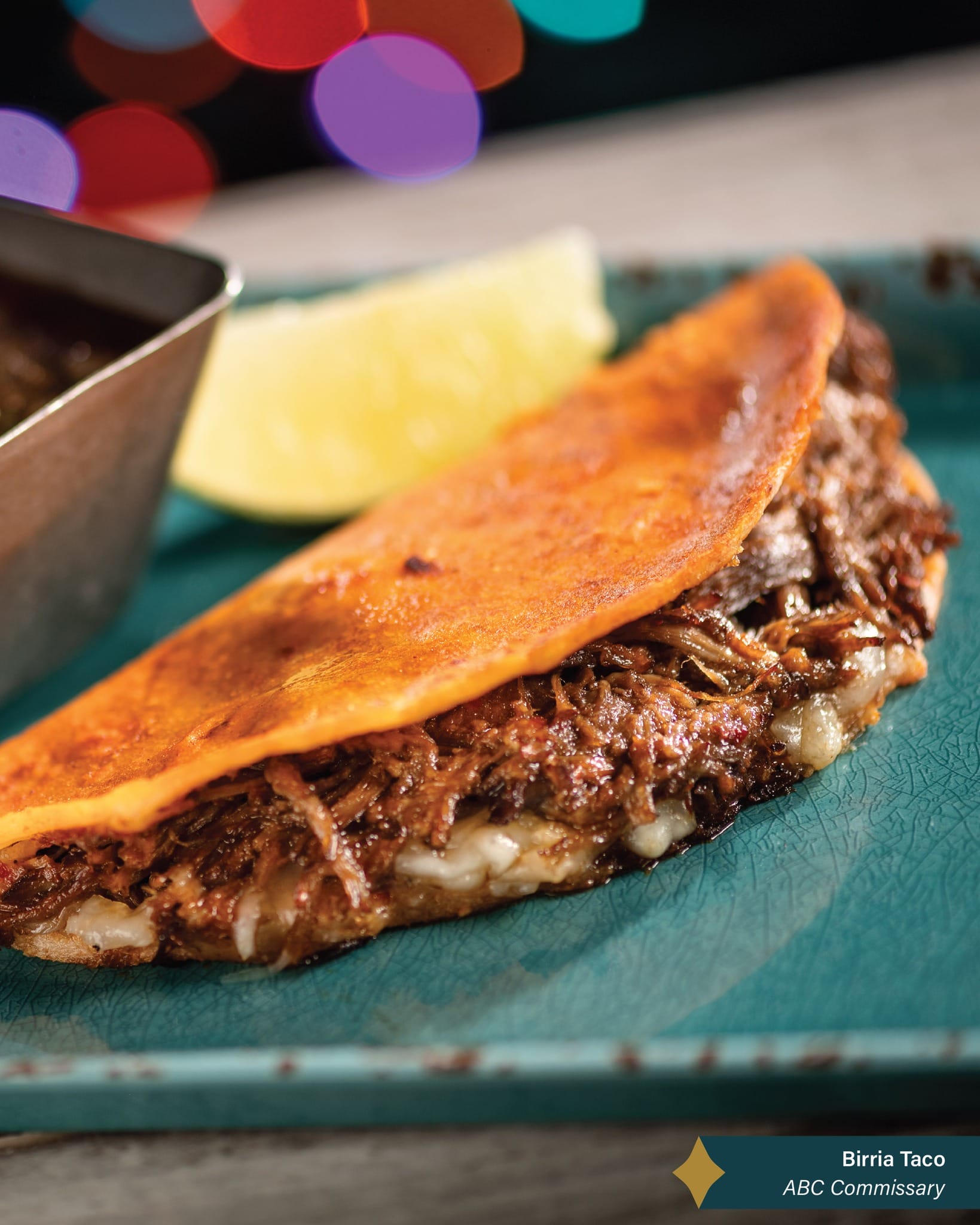 Close-up of a birria taco with shredded beef and melted cheese, served with a lemon wedge on a teal plate at Disneyland Resort.