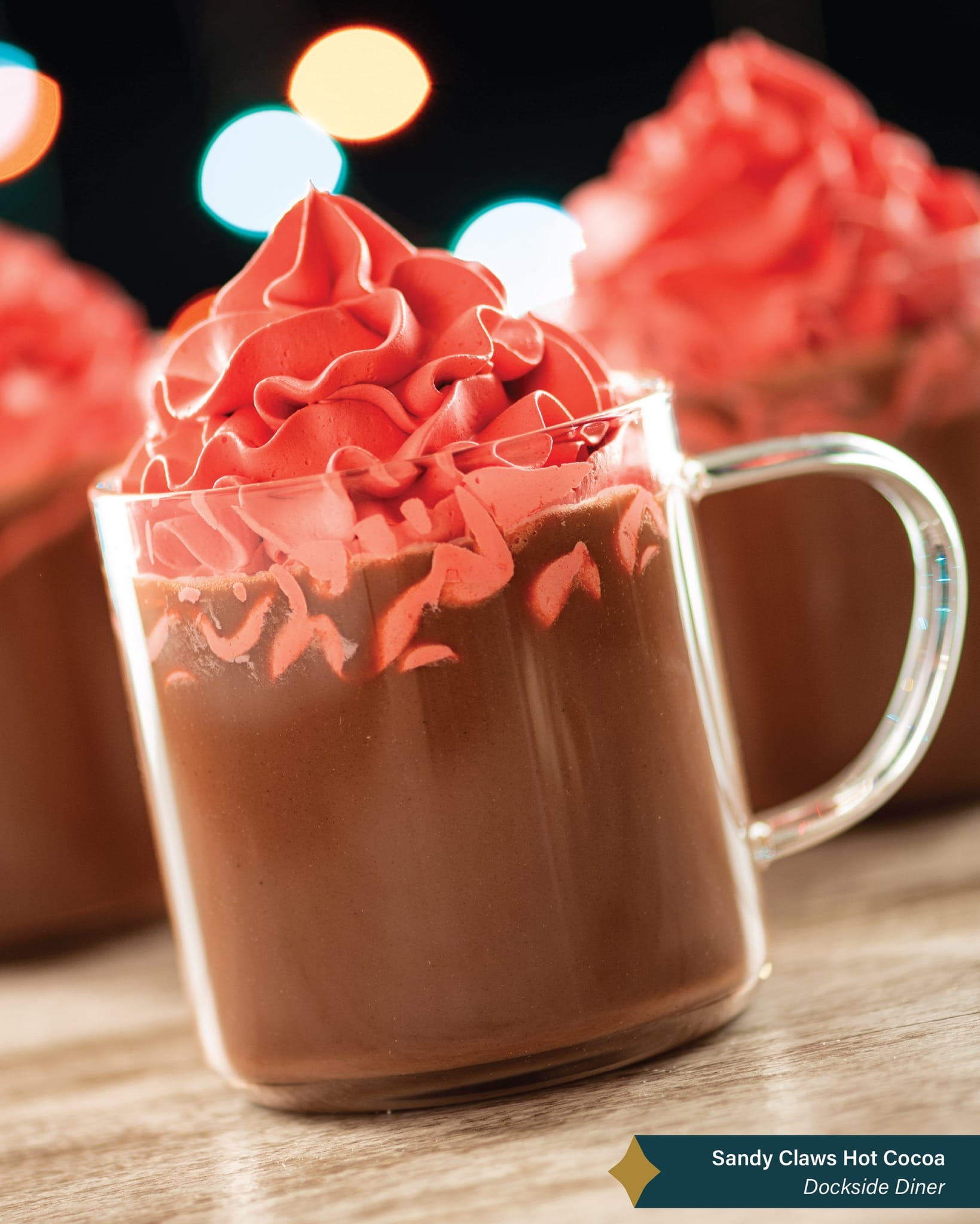 A clear mug of hot cocoa with bright red whipped cream, set against blurred festive lights reminiscent of Main Street at Disneyland.