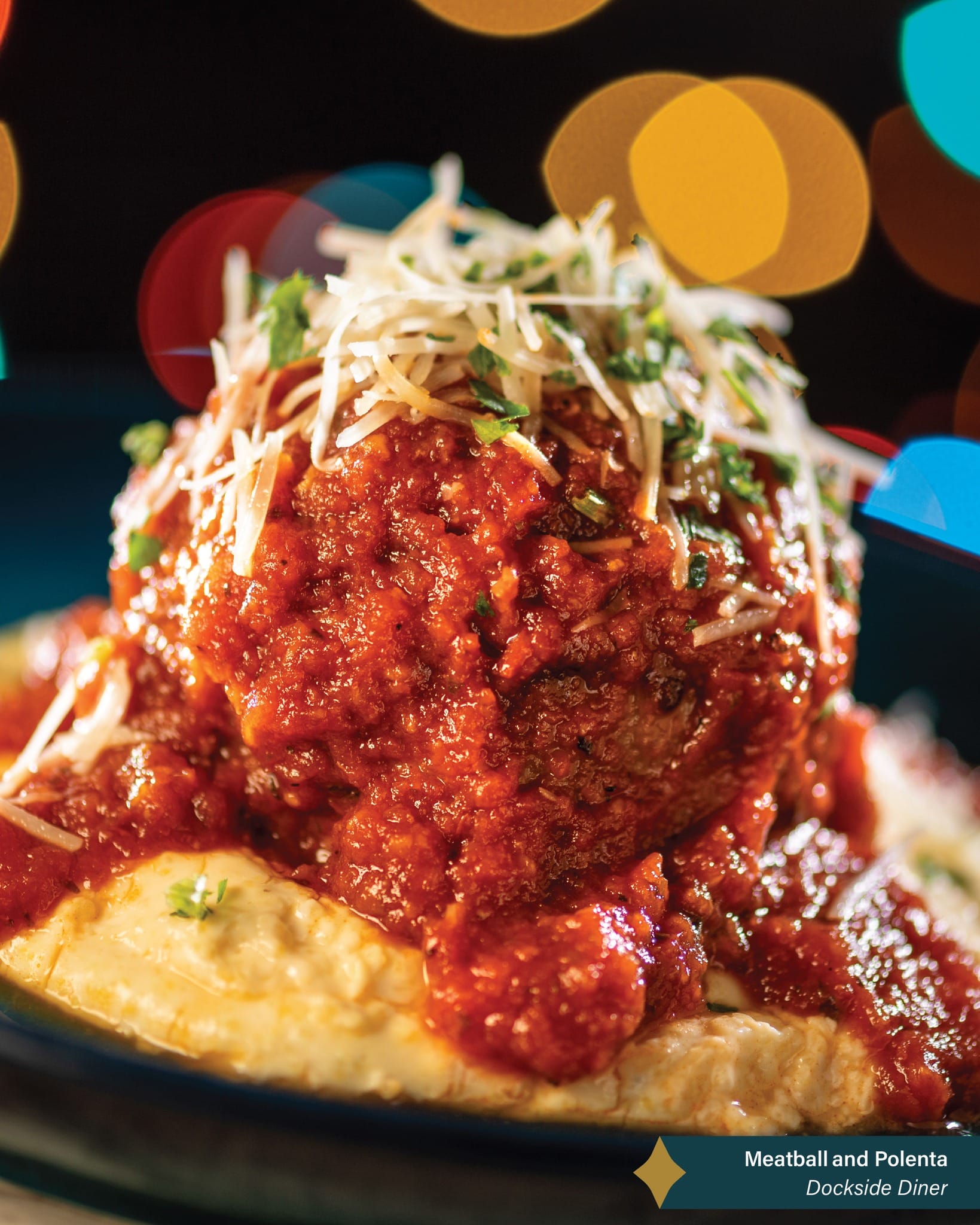 A meatball with marinara sauce and cheese on creamy polenta, with festive, Disney park-like bokeh lights in the background.