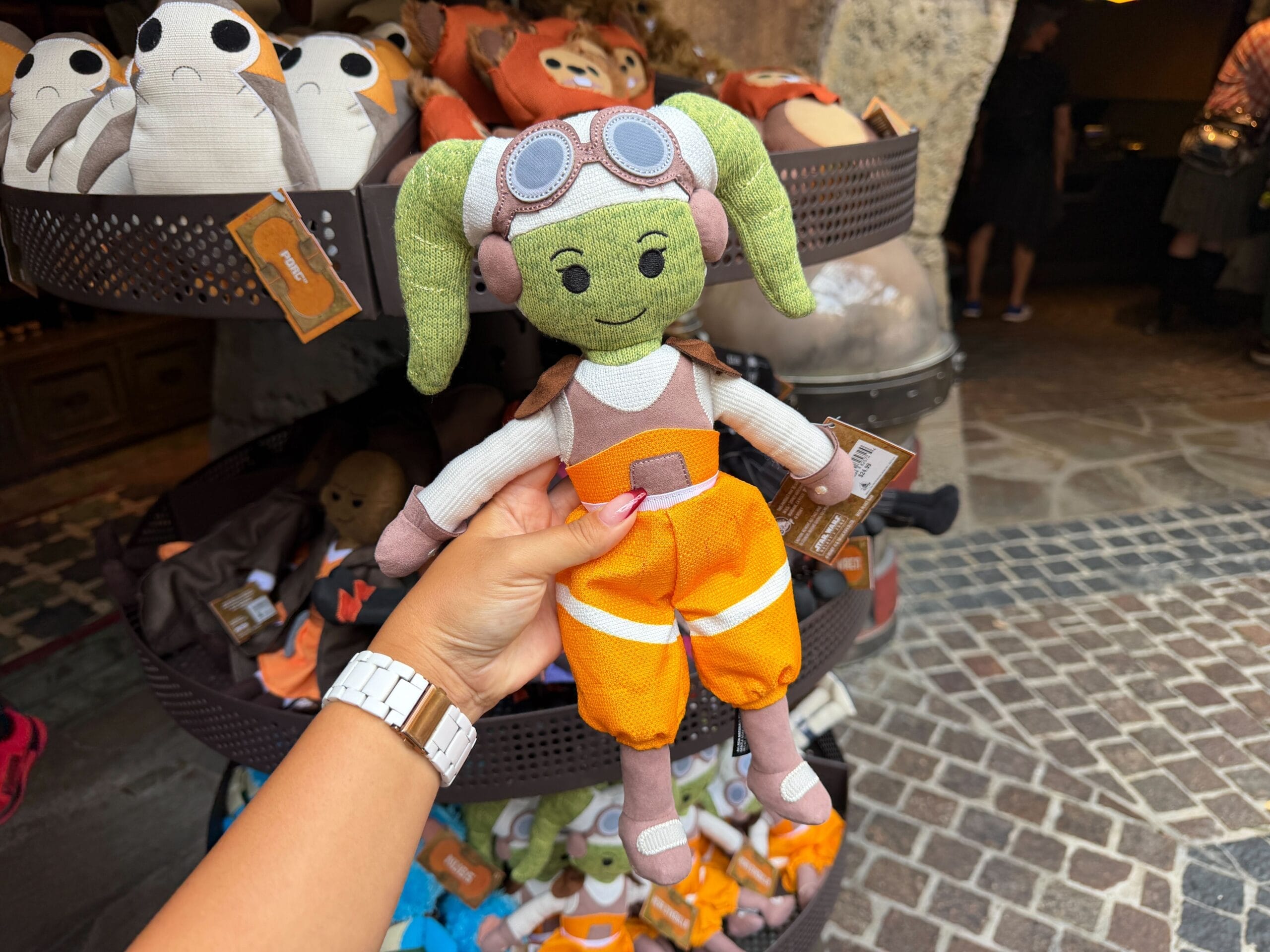 A hand holds a green plush toy in front of an Epic Universe display, reminiscent of Disney park souvenir stands near iconic castles.