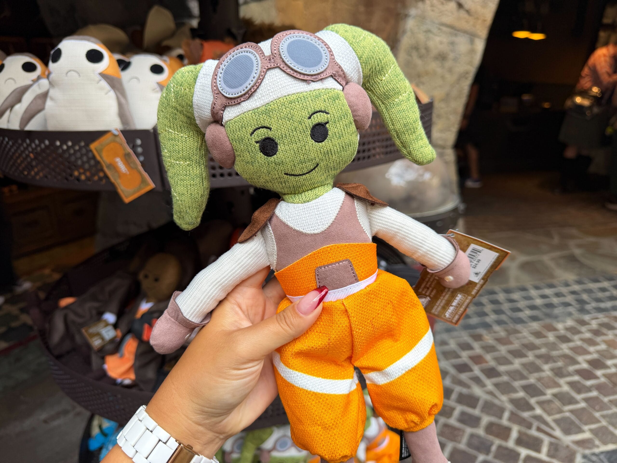 A hand holds a green Hera plush doll with goggles and orange pants at Galaxy's Edge, plushies fill the background.