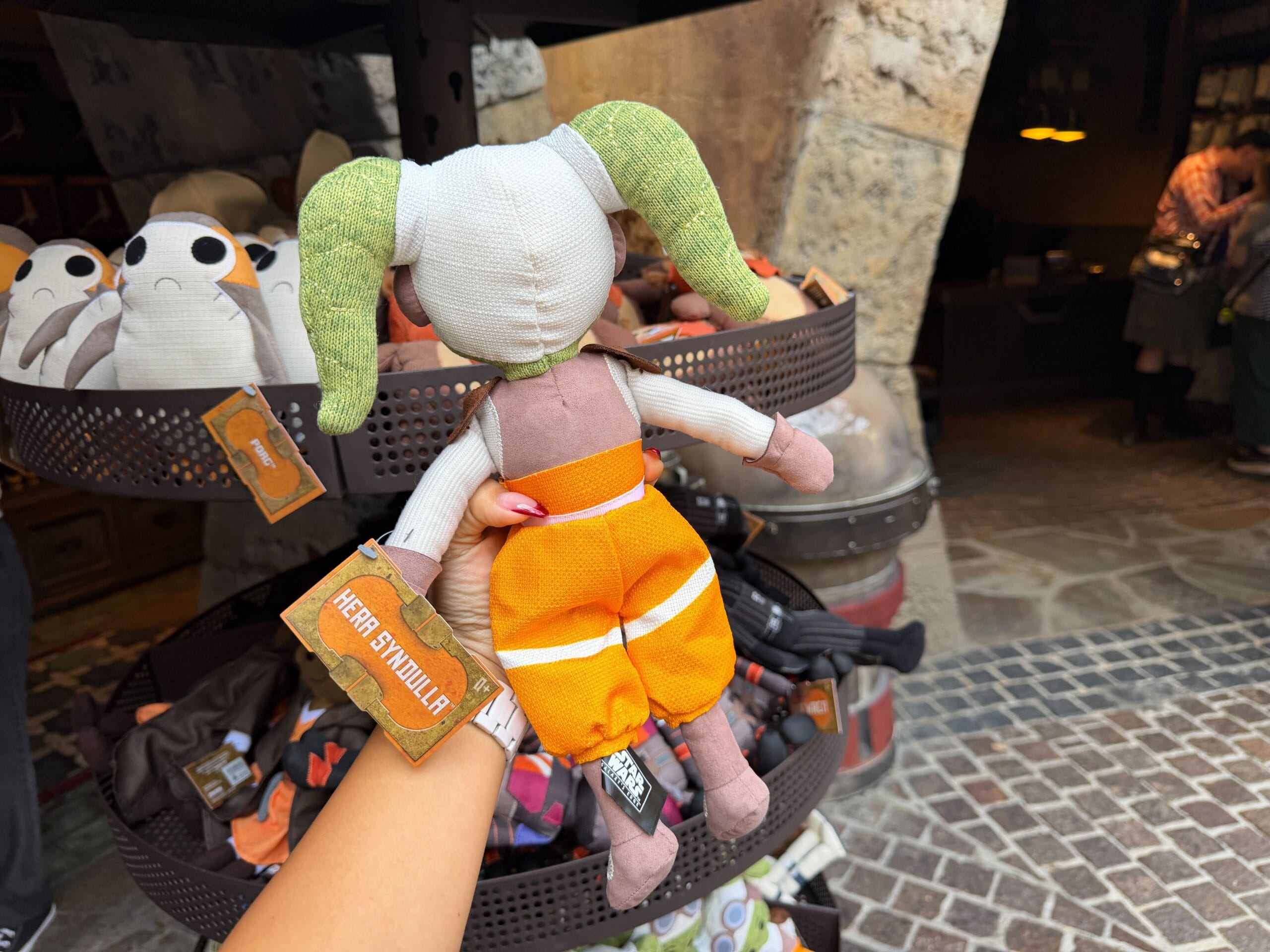 A hand holds a green and white Hera plush at a Disney park store display, surrounded by other themed plush toys.