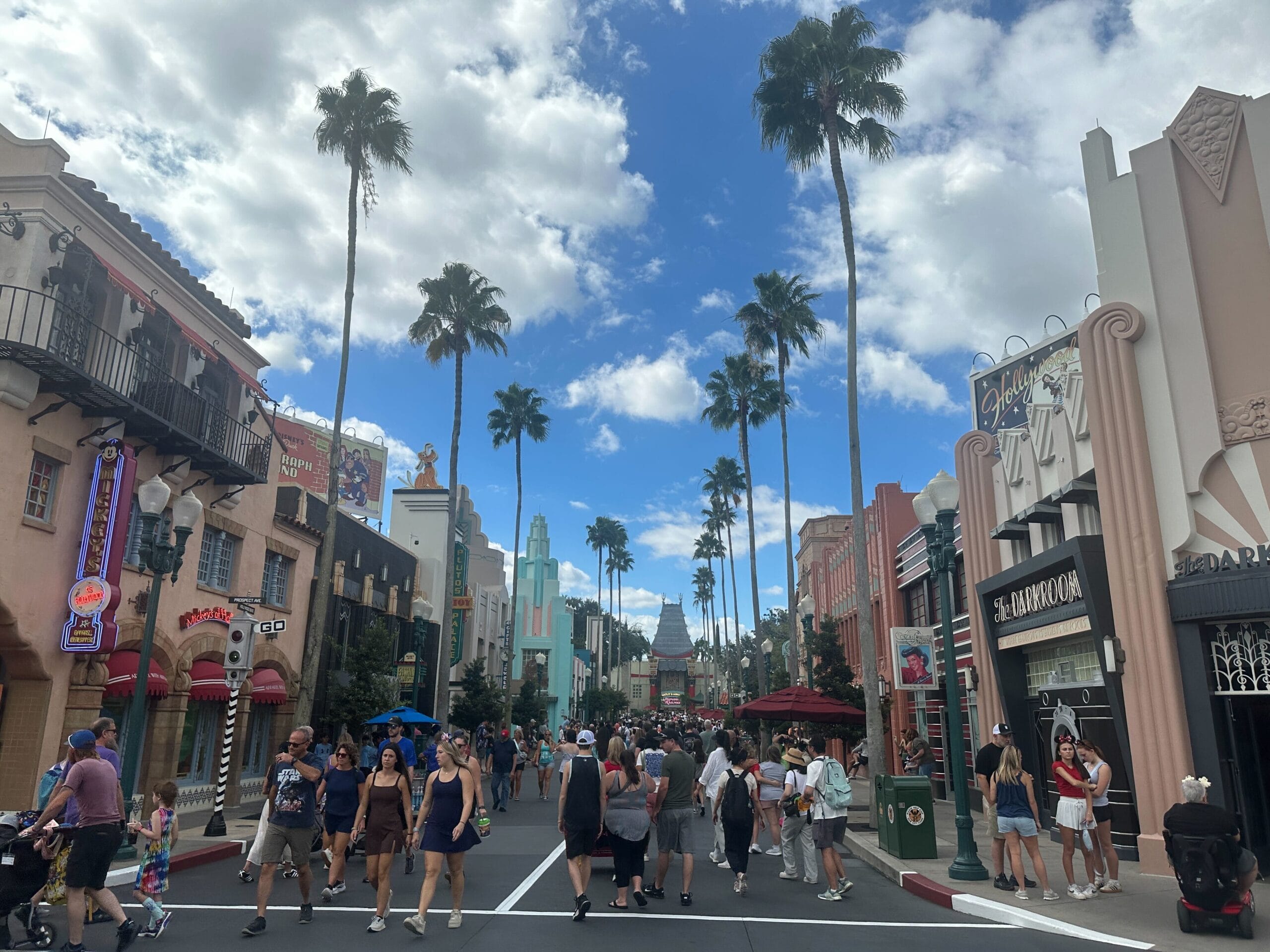 People walk along a palm-lined street with shops under a blue sky at Hollywood Studios, one of Walt Disney World's theme parks.