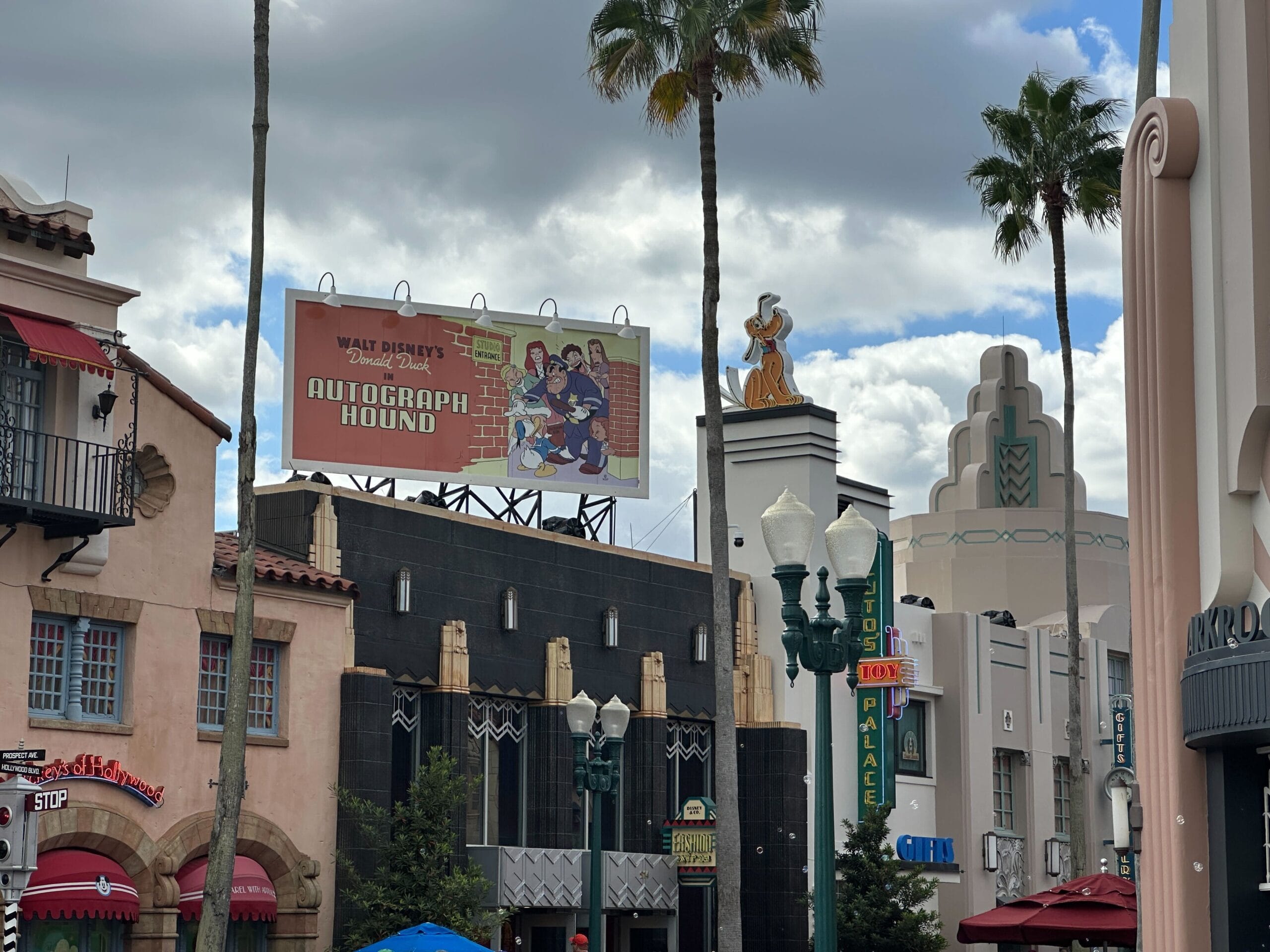 A festive Hollywood Studios street with “Autograph Hound” billboard and art deco buildings, lit up for the holidays. No castle here!