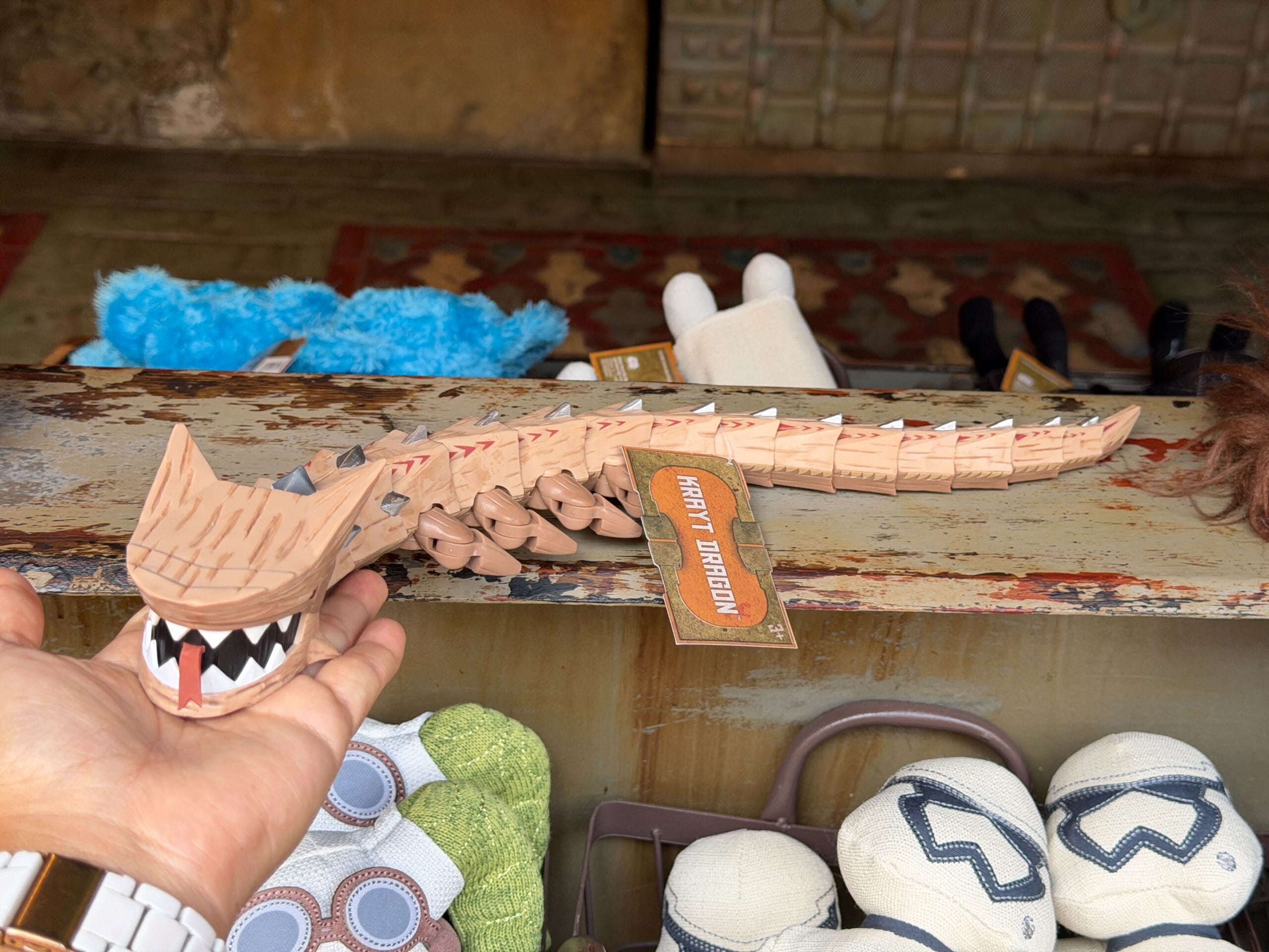 A hand displays a wooden, jointed Krayt Dragon toy above plushies on a rustic table